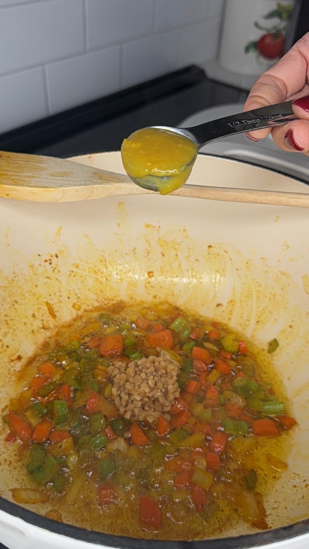 A hand holds a teaspoon of yellow paste above a pot with diced carrots, peppers, and seasonings, ready to add the paste to the mixture. A wooden spoon rests on the pot edge.