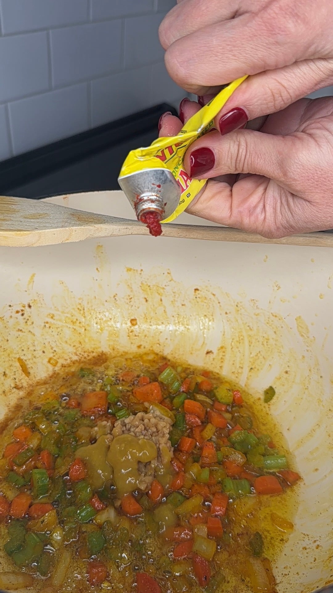 A hand squeezes tomato paste from a tube into a bowl of chopped vegetables and sauce, preparing a dish. The bowl contains carrots, green peppers, and a brown sauce.