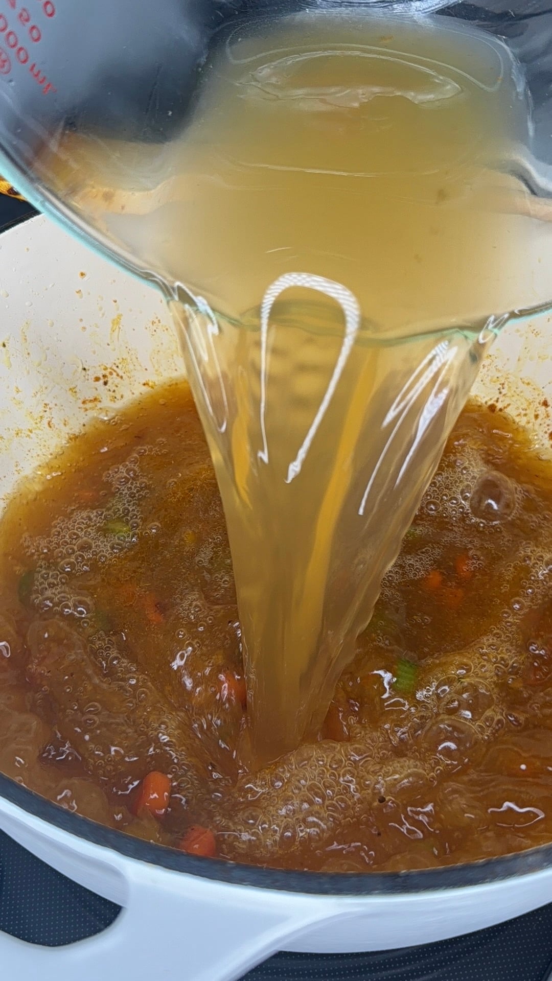 Broth is being poured from a glass measuring cup into a white pot filled with a simmering mixture of vegetables and seasonings.