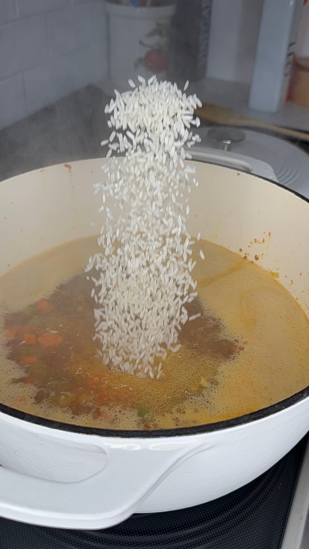 A stream of uncooked rice is being poured into a large white pot filled with simmering broth and vegetables on a stove.