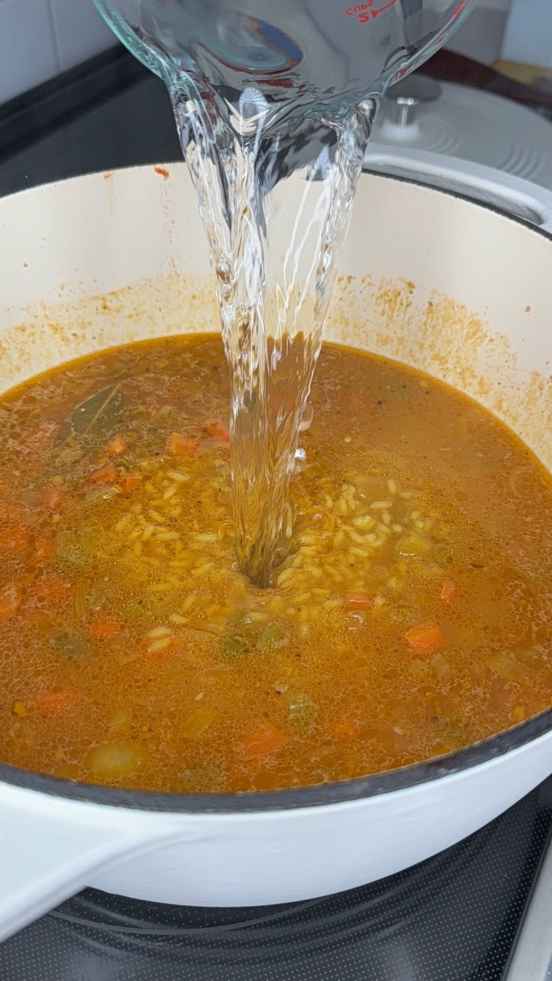Water is being poured from a glass measuring cup into a pot of soup with rice, vegetables, and spices, simmering on a stovetop.