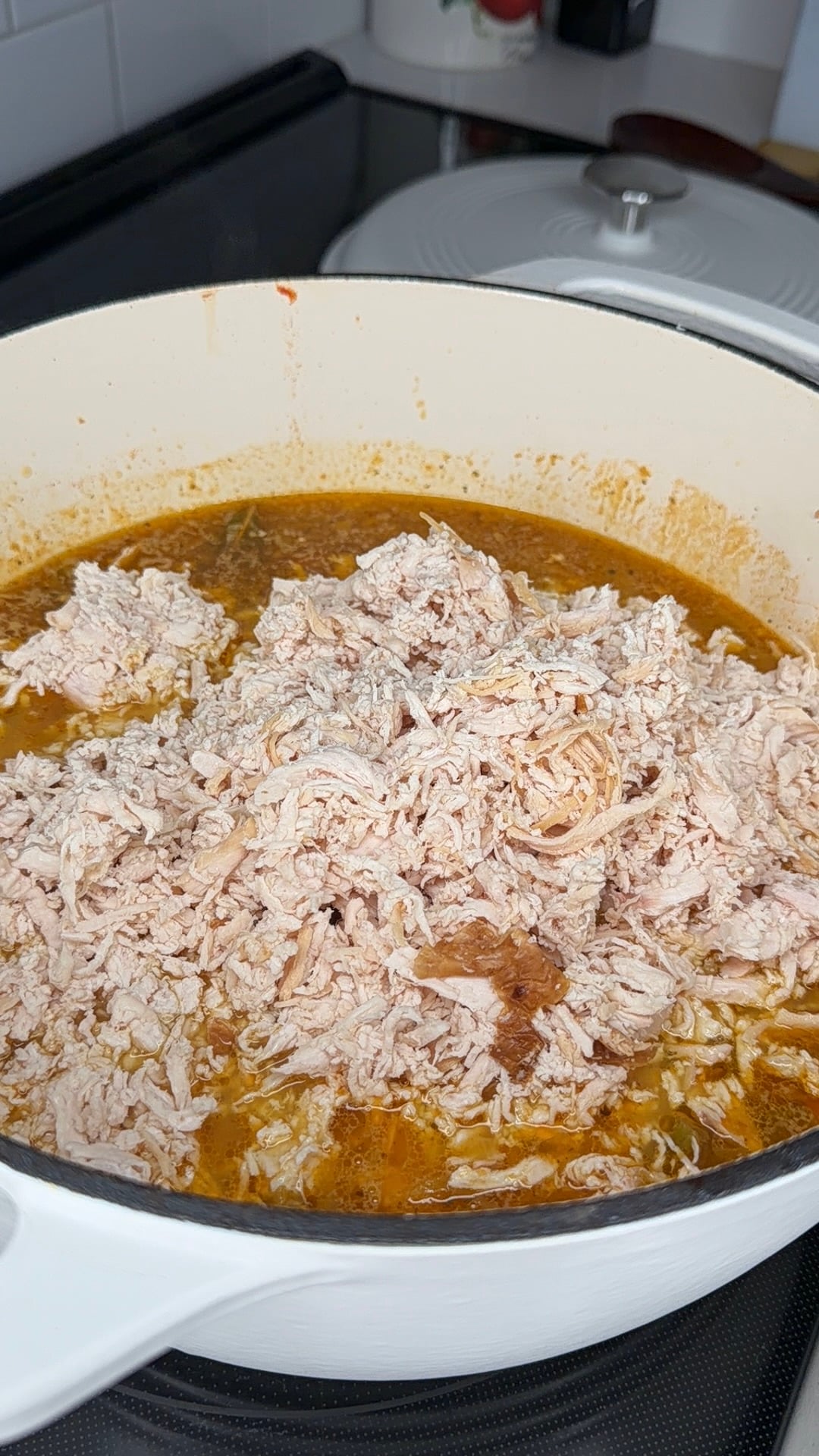 Shredded chicken sits in a white pot filled with orange broth and spices, cooking on a stovetop. The pot lid rests in the background.