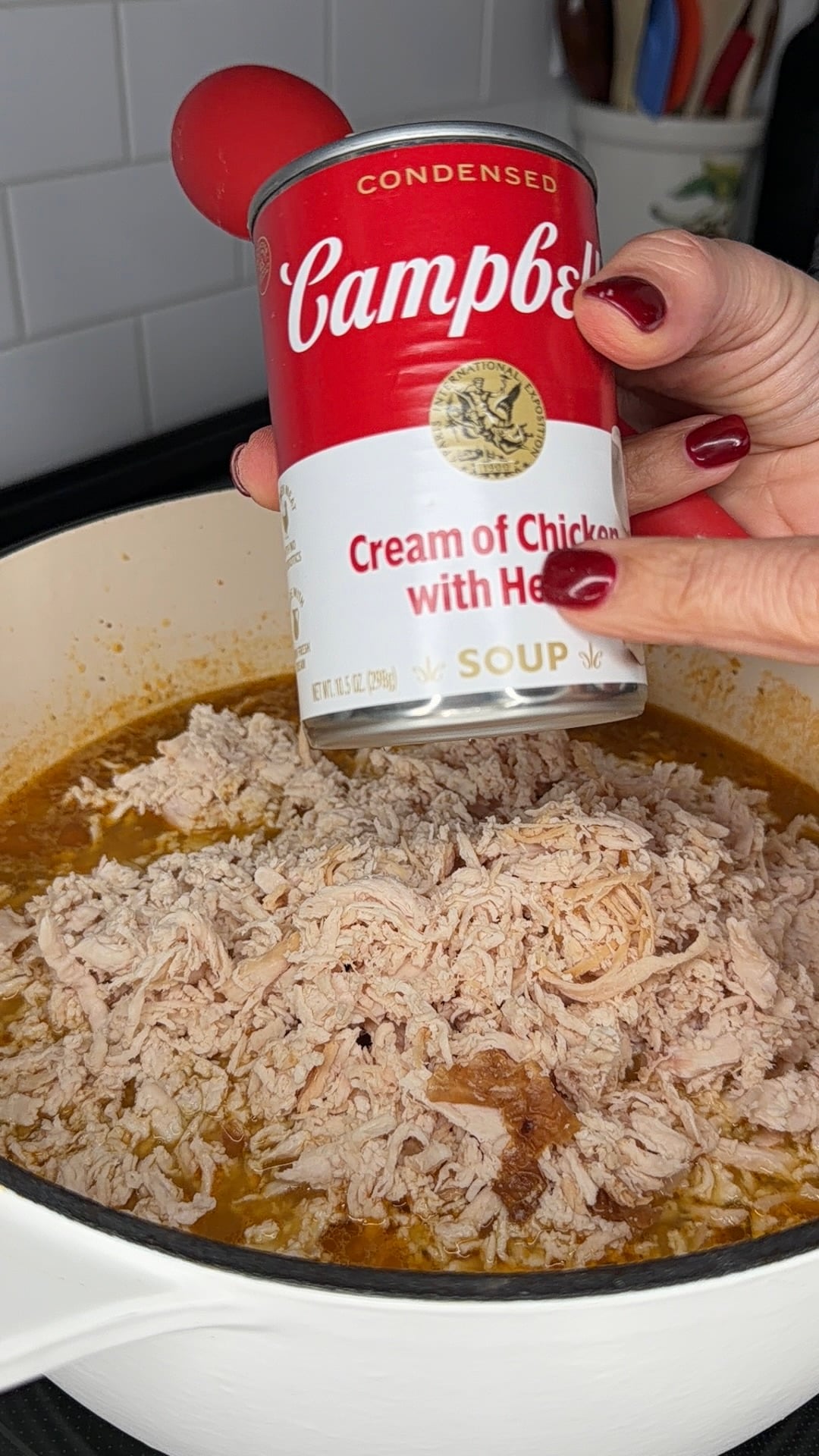 A hand holds a can of Campbells Cream of Chicken with Herbs soup over a pot filled with shredded chicken in broth on a stove.