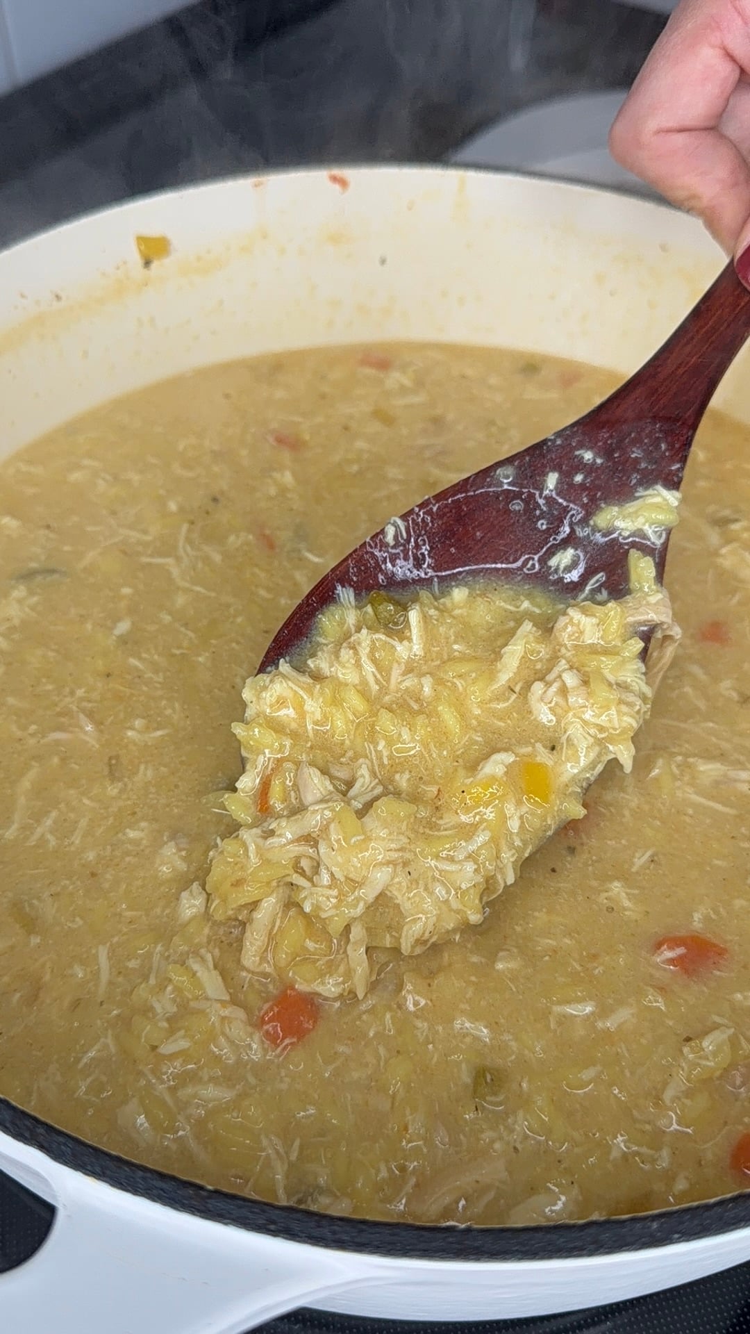 A wooden spoon scoops up a thick, creamy chicken and rice soup with vegetables from a white pot on a stove.