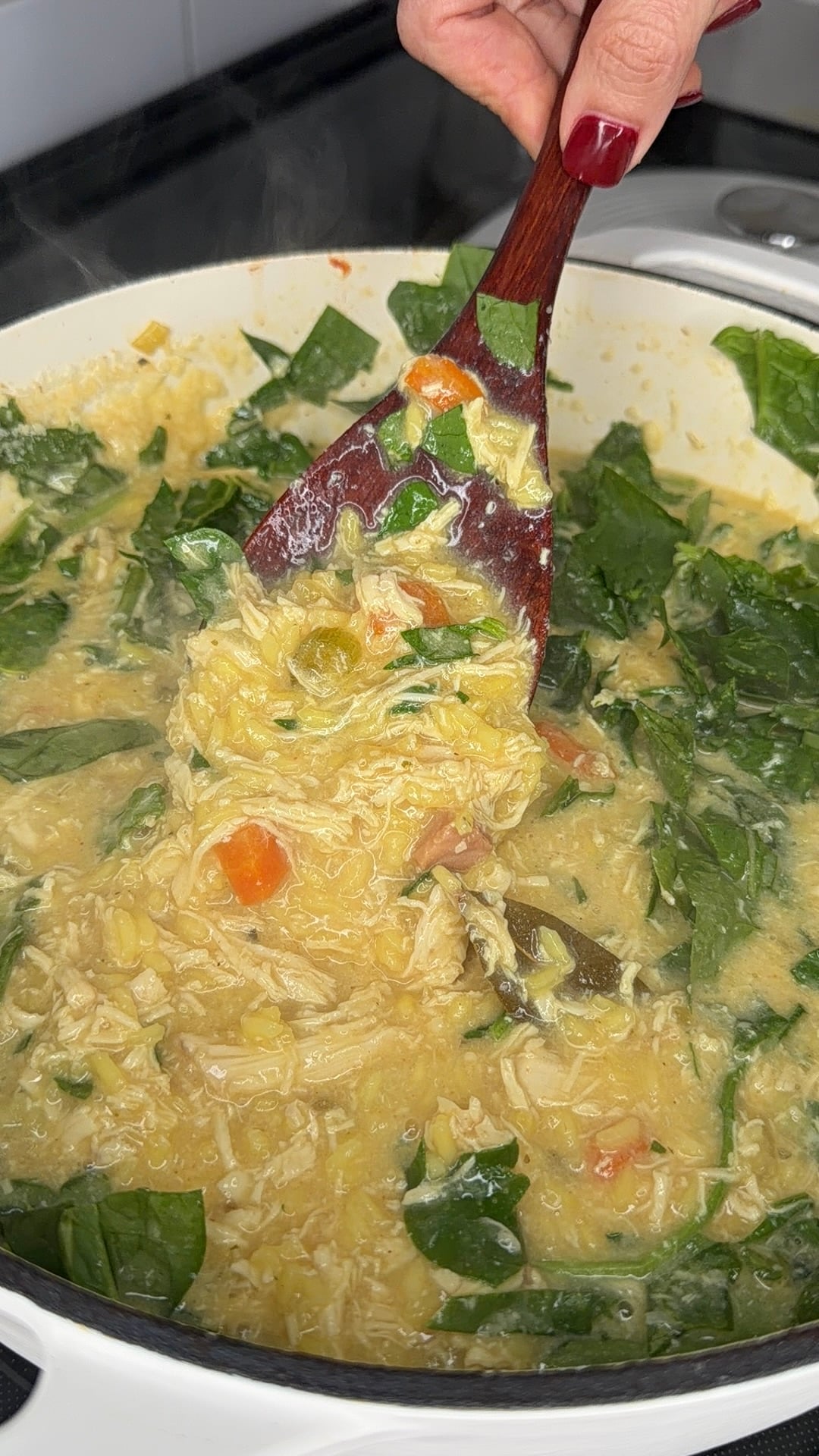A hand with red nails stirs a pot of creamy chicken soup with spinach, carrots, and orzo, using a wooden spoon. Steam rises from the pot.
