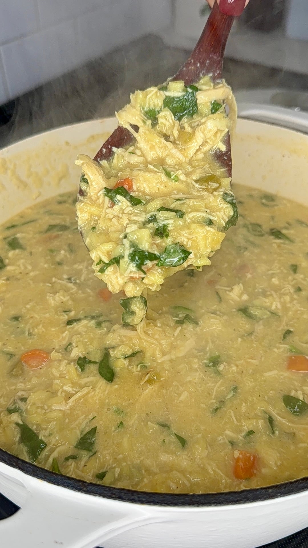 A wooden spoon lifts a serving of thick, hearty chicken soup with rice, carrots, and spinach from a white pot on a stove. Steam rises from the hot soup.