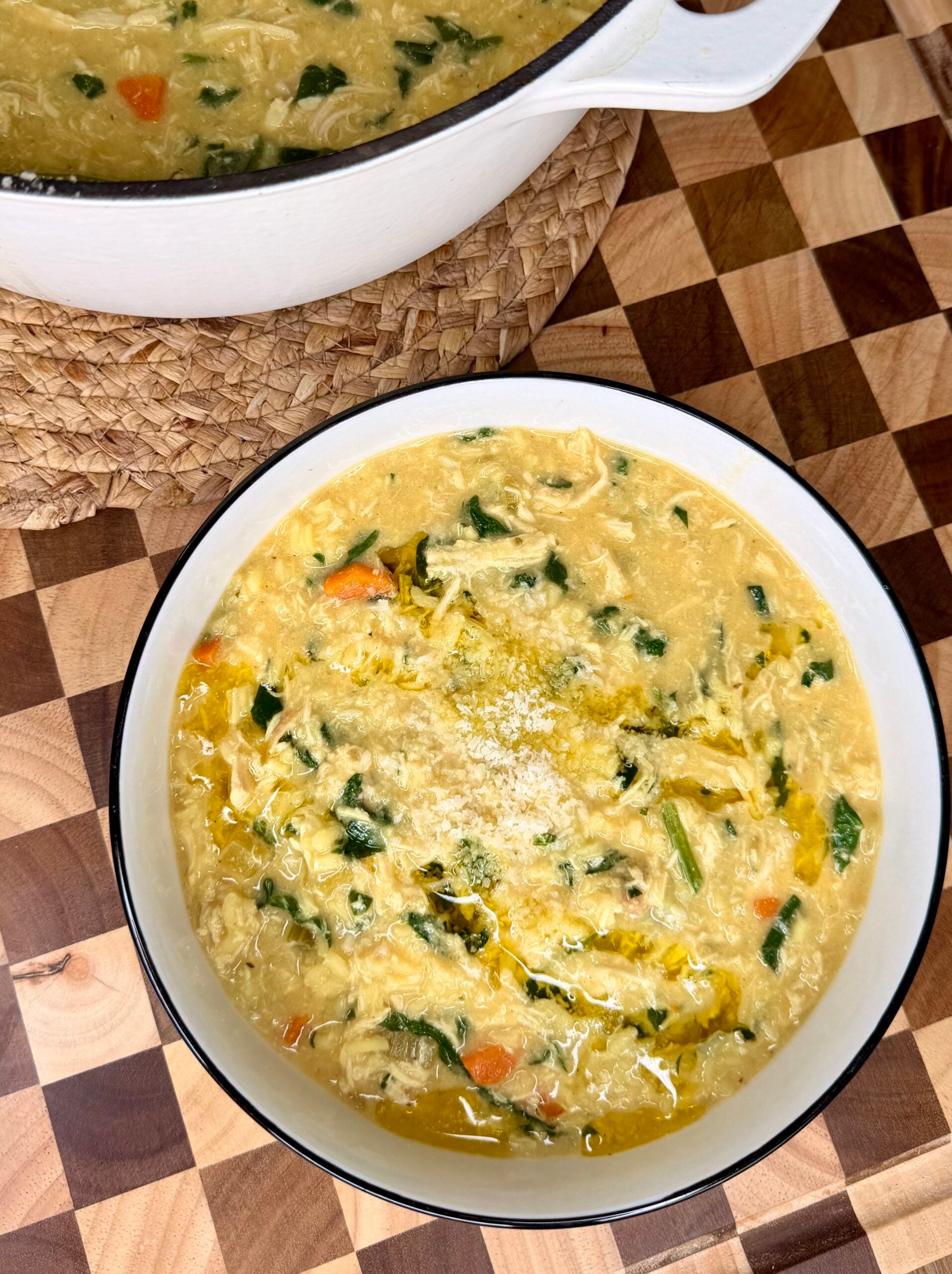 A bowl of chicken soup with shredded chicken, carrots, spinach, and broth sits on a checkered wooden surface next to a pot of more soup.