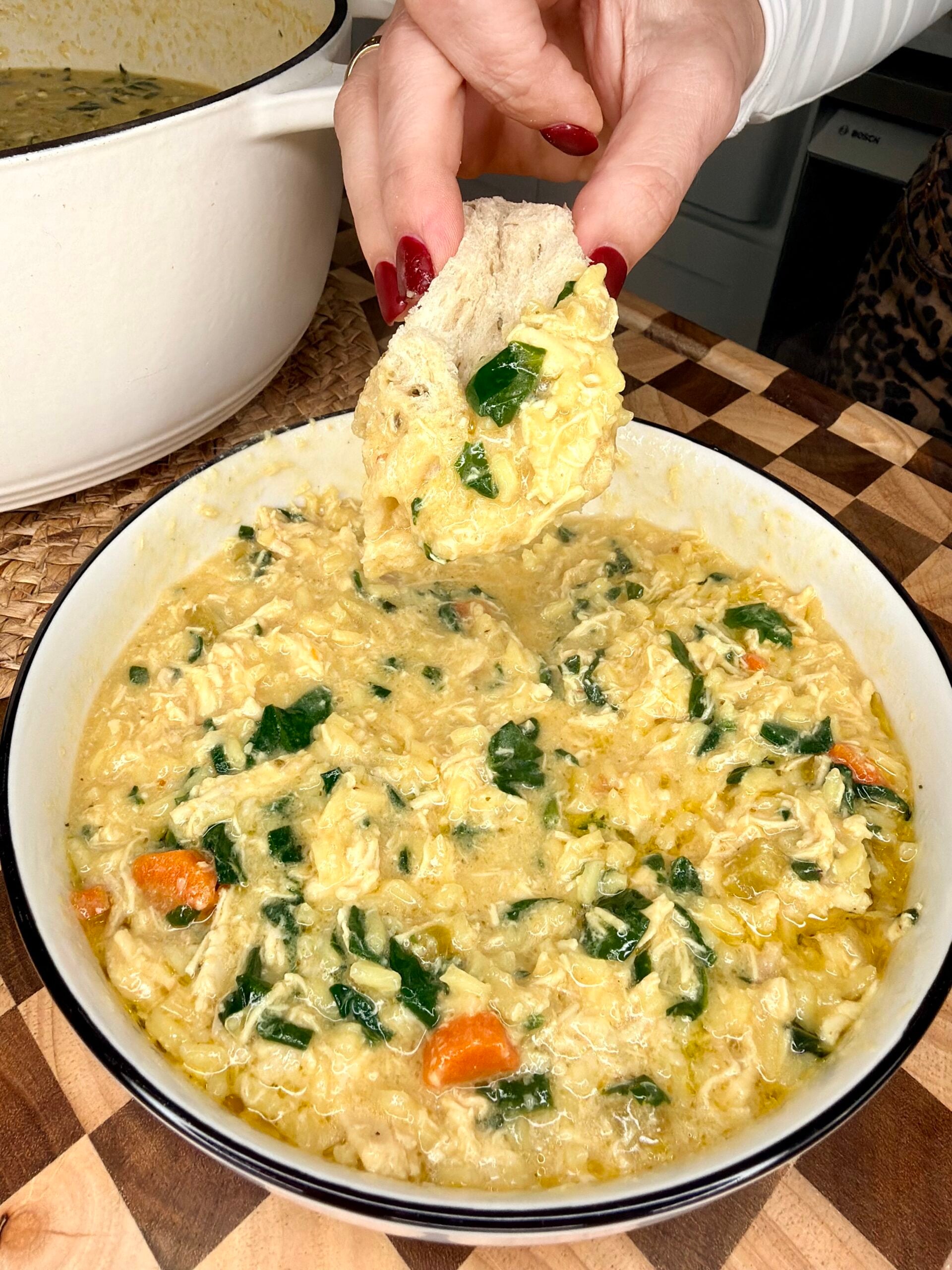 A hand with red nail polish dips bread into a bowl of creamy soup with shredded chicken, rice, spinach, and carrots on a checkered wooden surface.