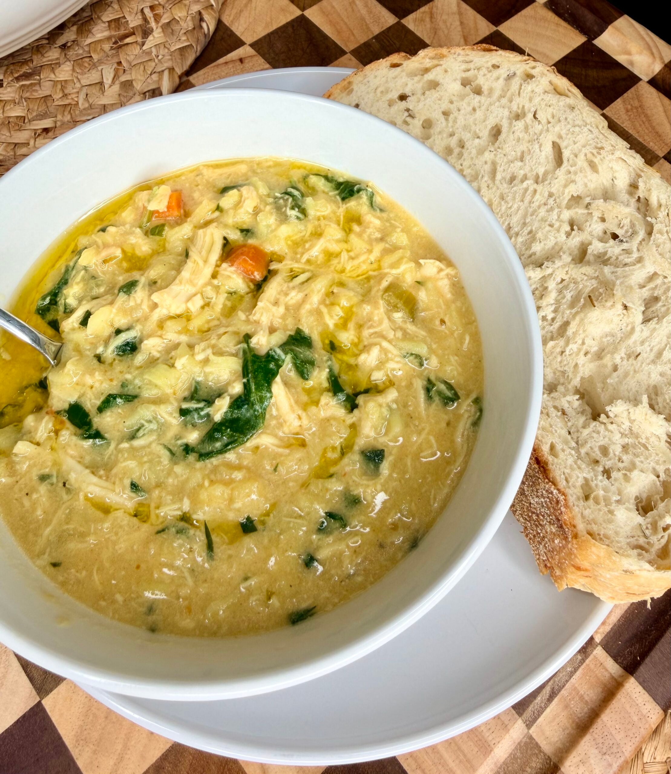 A bowl of creamy chicken soup with vegetables and spinach, served with two slices of rustic bread on a white plate, set on a wooden checkered surface.