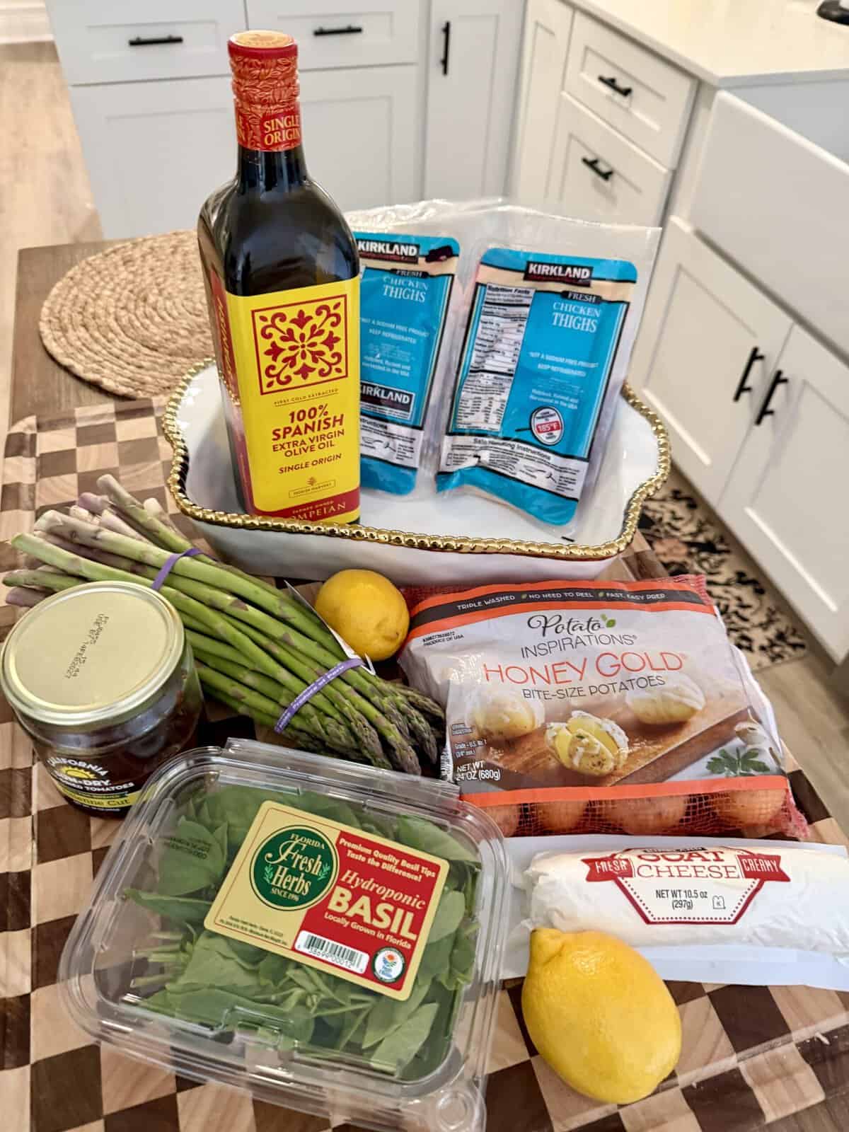 A tray holds olive oil and chicken thighs, surrounded by asparagus, basil, a lemon, honey gold potatoes, goat cheese, and a jar of pesto, all set on a kitchen countertop.