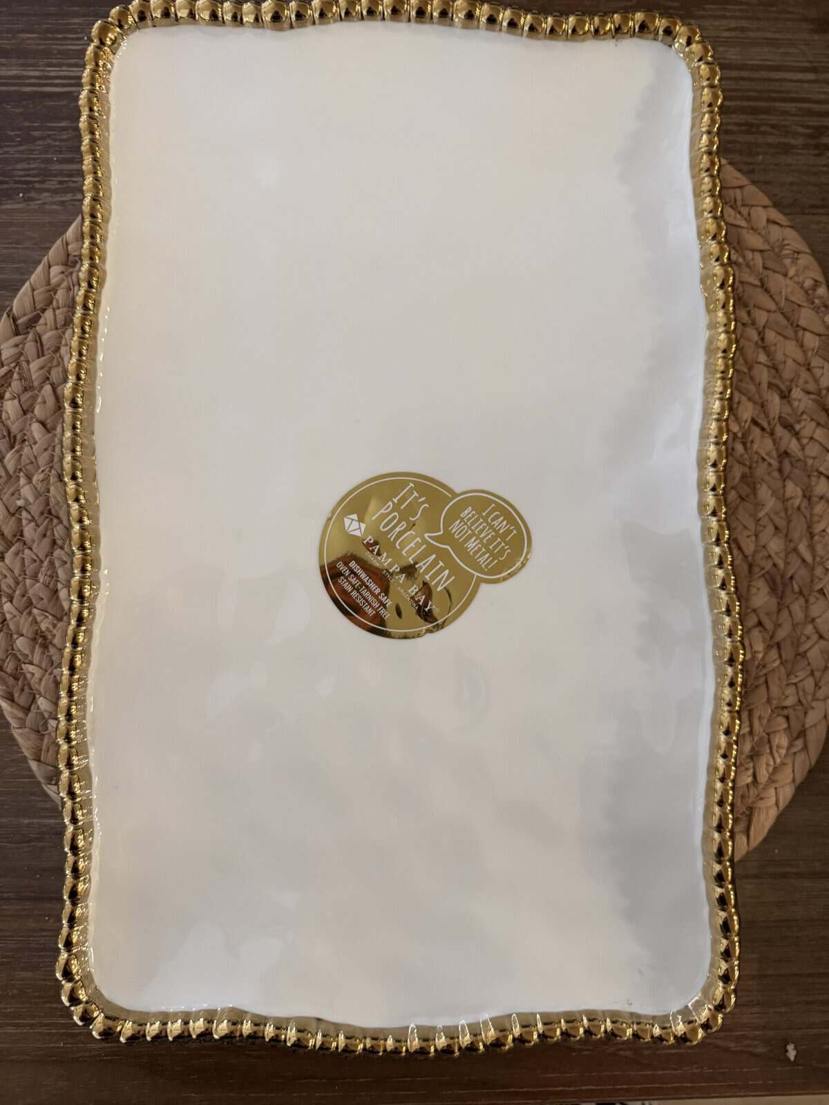A rectangular white porcelain platter with a wavy gold beaded edge, placed on a woven placemat. There is a round gold sticker in the center of the platter.