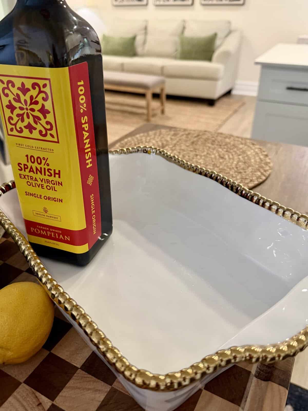 A bottle of Pompeian 100% Spanish extra virgin olive oil sits next to a lemon and a white serving dish with a gold beaded edge on a kitchen counter. A sofa and table are visible in the background.