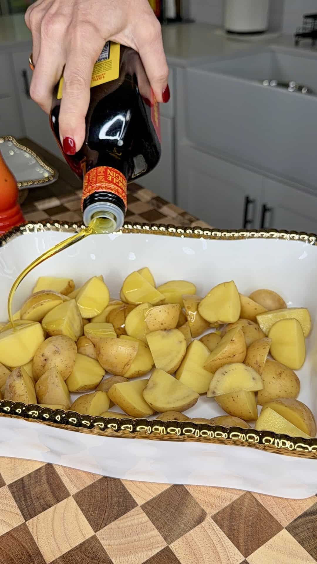 A hand pours olive oil over chopped yellow potatoes in a white, gold-rimmed dish on a wooden countertop in a kitchen.