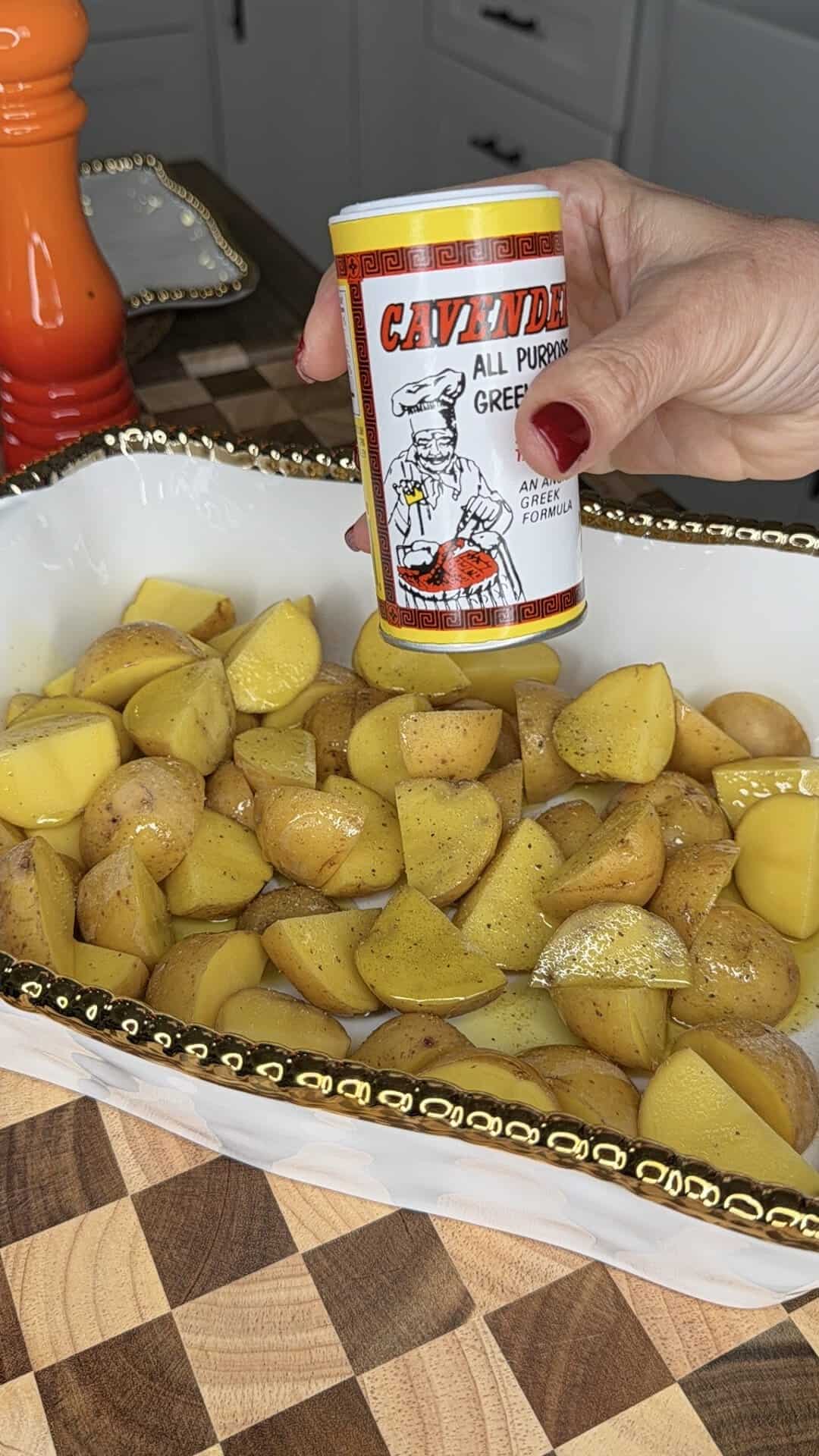 A hand sprinkles Cavenders All Purpose Greek Seasoning over a white dish filled with chopped yellow potatoes on a kitchen counter.