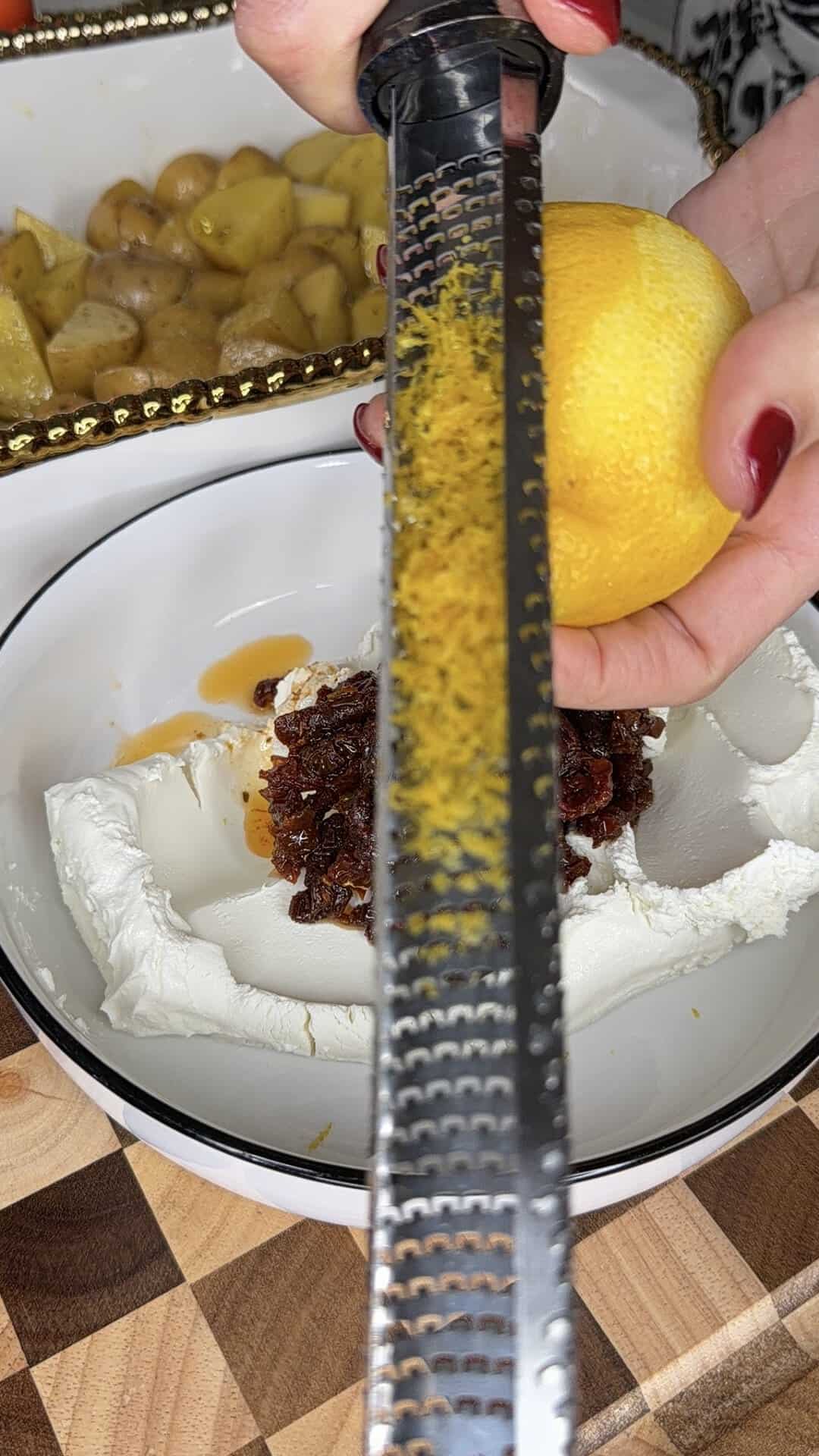 A hand grates lemon zest over a bowl with cream cheese and chopped dates. A tray of roasted potatoes sits in the background on a wooden cutting board.