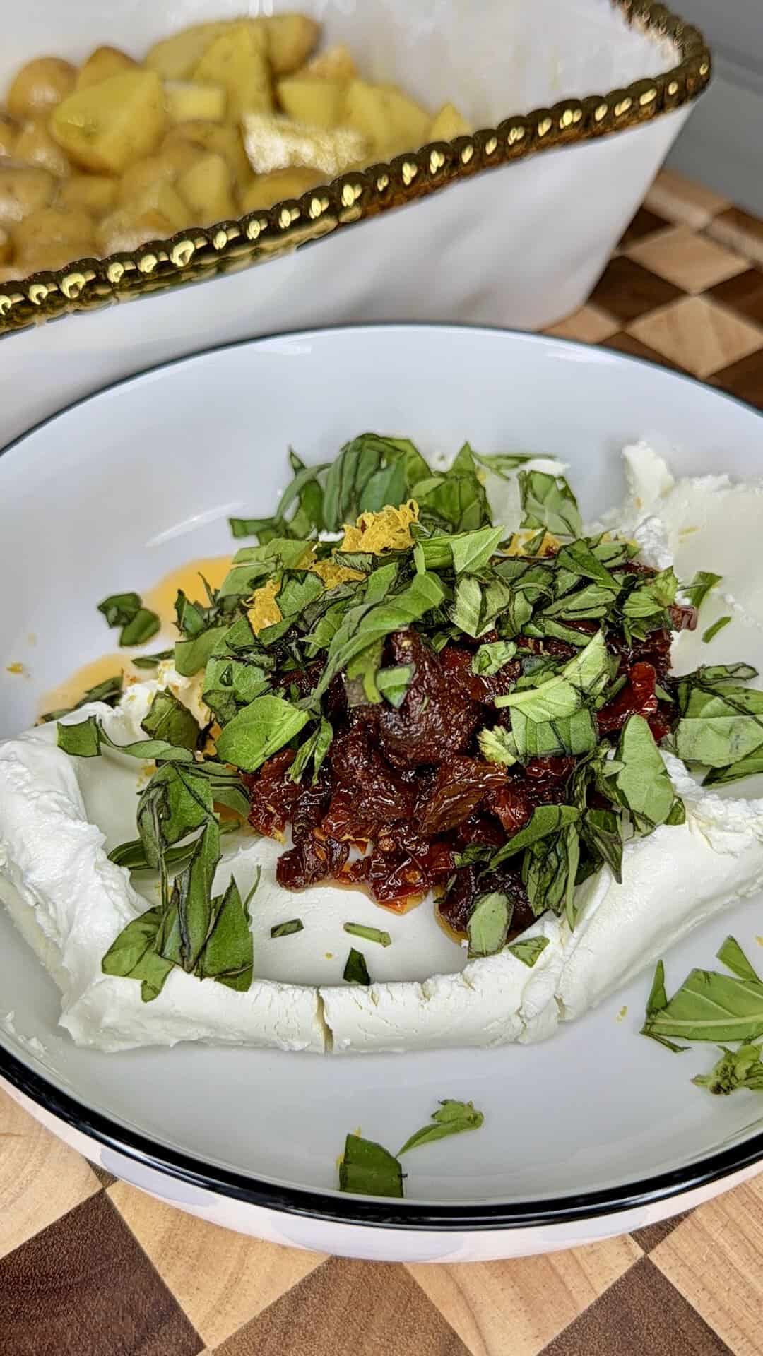 A bowl with whipped ricotta, topped with chopped fresh herbs, sun-dried tomatoes, and orange zest. In the background, a dish of roasted potatoes is partially visible.
