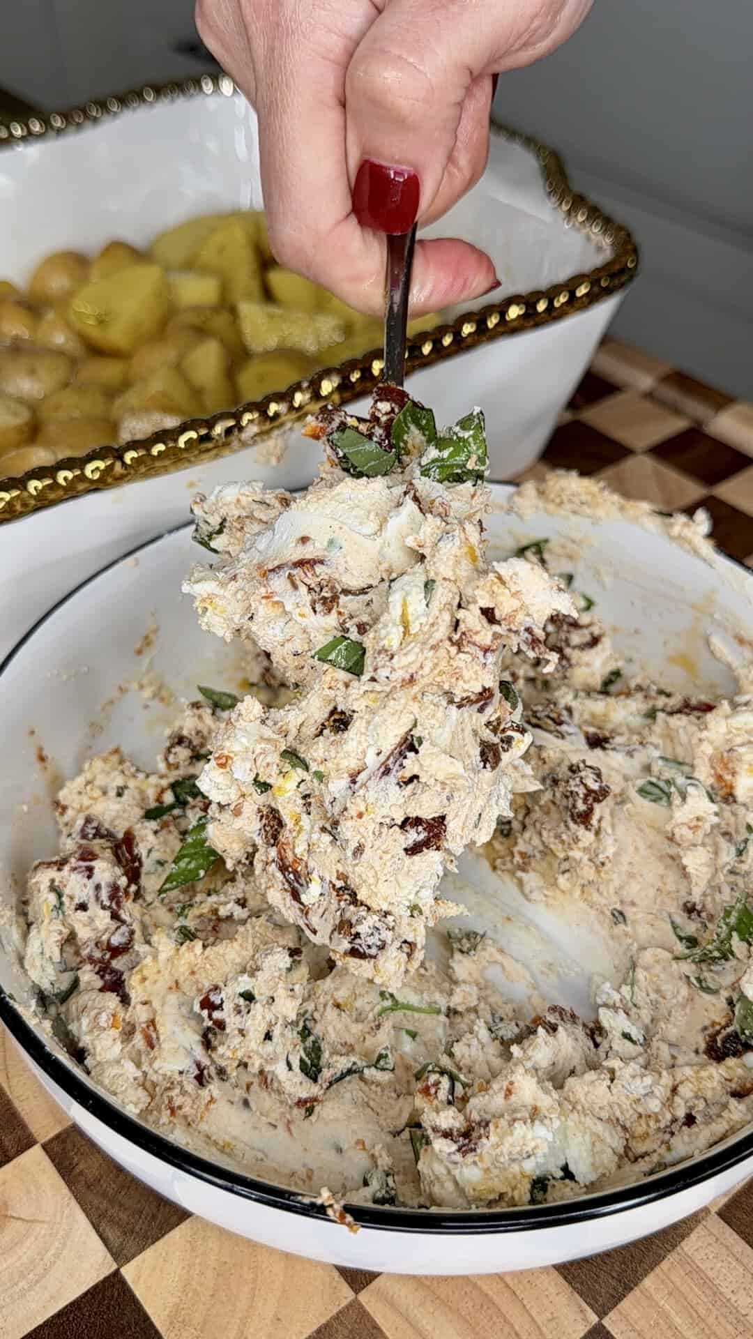 A hand scoops a chunky, herbed cheese mixture from a white bowl. In the background, a dish with roasted potatoes sits on a checkered wooden surface.