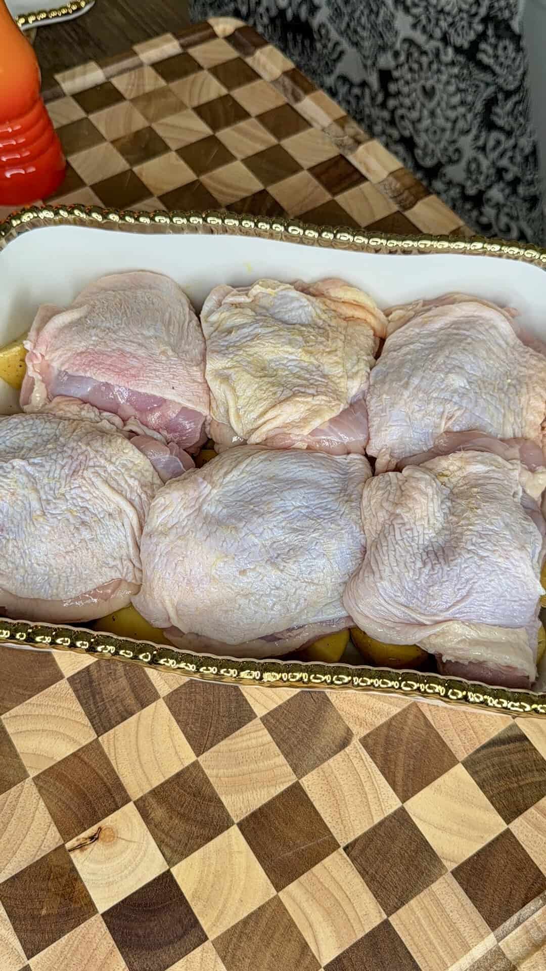 Six raw, skin-on chicken thighs arranged in a white baking dish with a gold trim, placed on a checkered wooden cutting board.