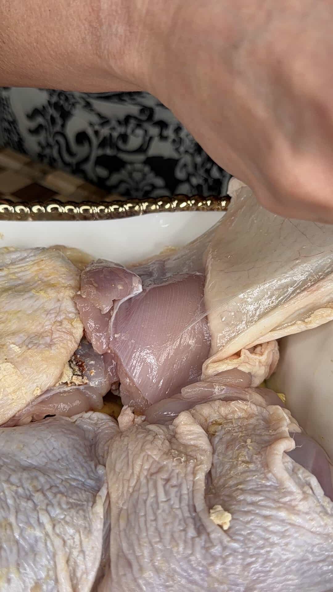 A hand lifts the skin off a raw chicken thigh in a white tray, preparing the meat for cooking.