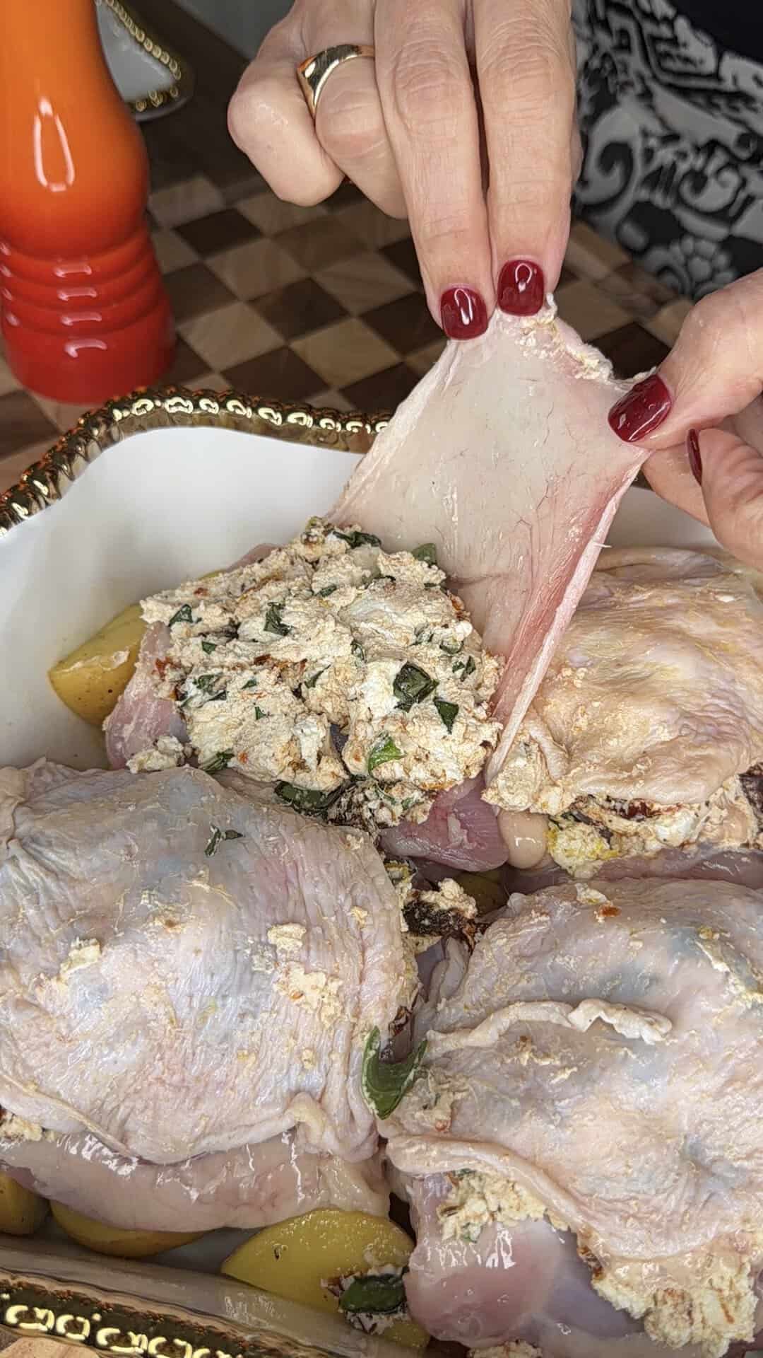A person stuffs seasoned mixture under the skin of raw chicken thighs in a baking dish with potato wedges, preparing the meat for cooking.