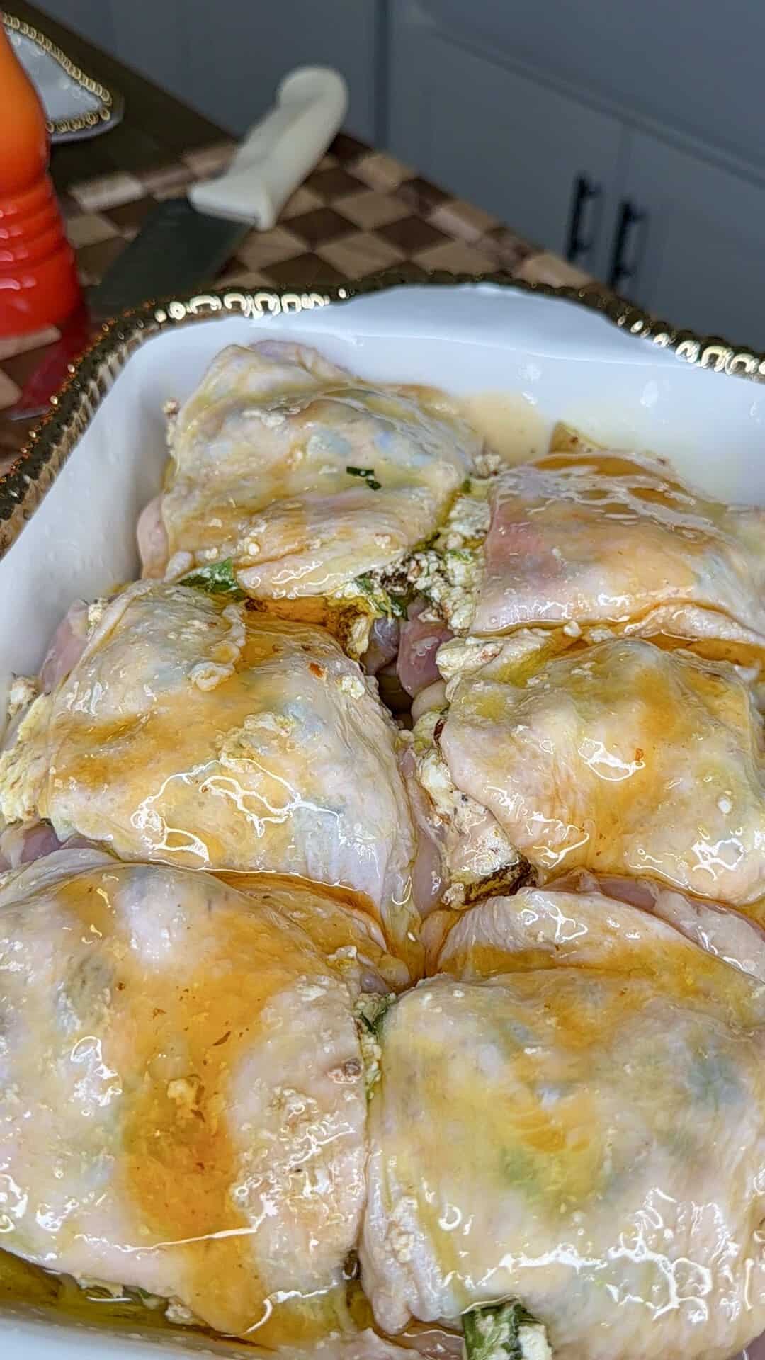 Raw seasoned chicken thighs stuffed with a creamy filling, arranged in a white dish and coated with a glaze, ready to be baked. The dish sits on a counter with a checkerboard pattern.