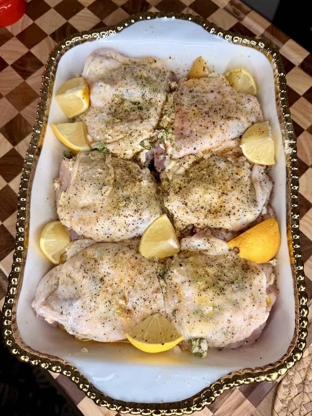 Raw, seasoned chicken thighs are arranged in a white, gold-rimmed baking dish with lemon wedges on a wooden cutting board. The chicken is sprinkled with black pepper and herbs, ready to be cooked.
