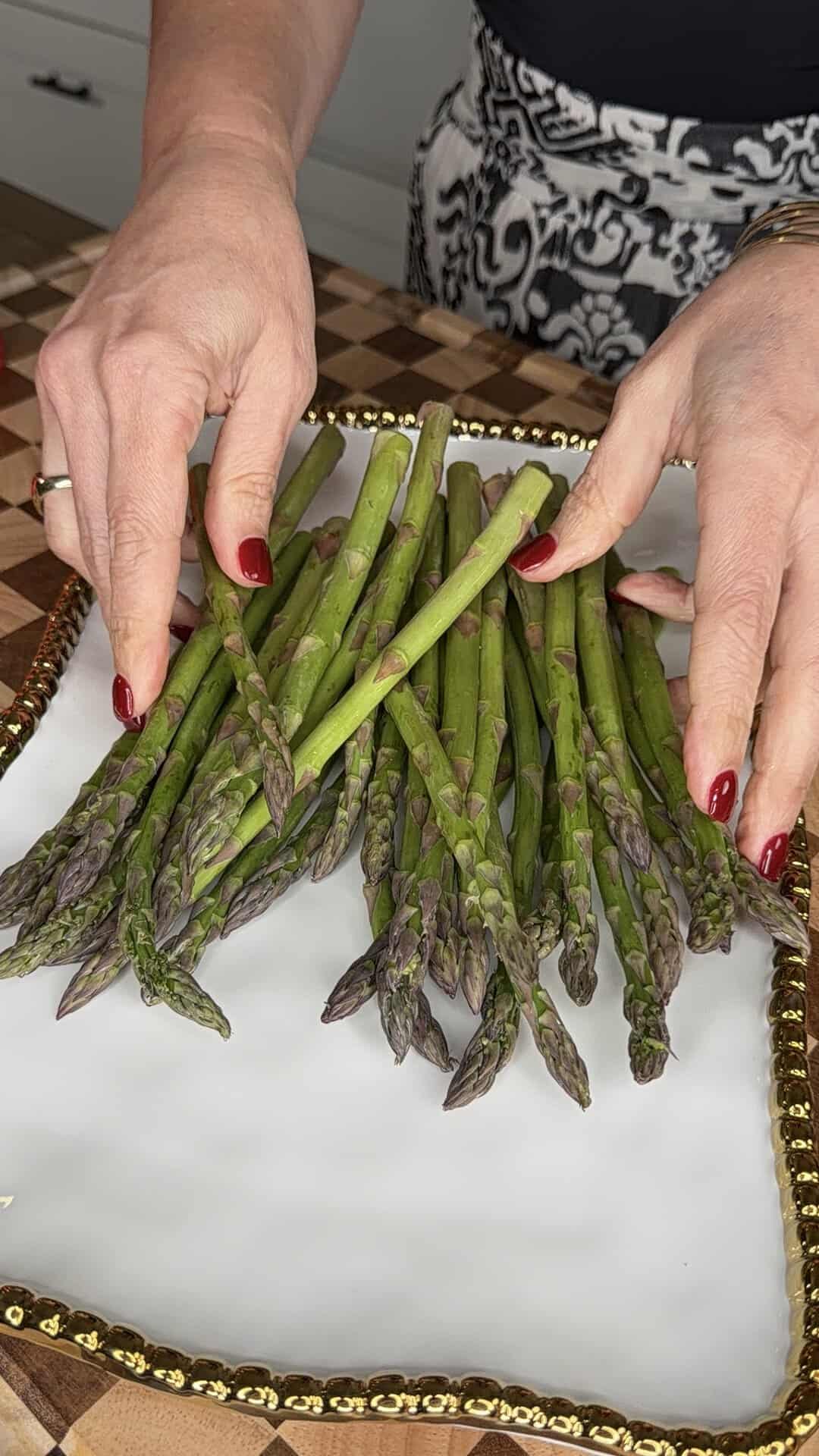 A person with red nail polish arranges fresh asparagus spears on a white, gold-rimmed plate set on a checkered wooden surface.