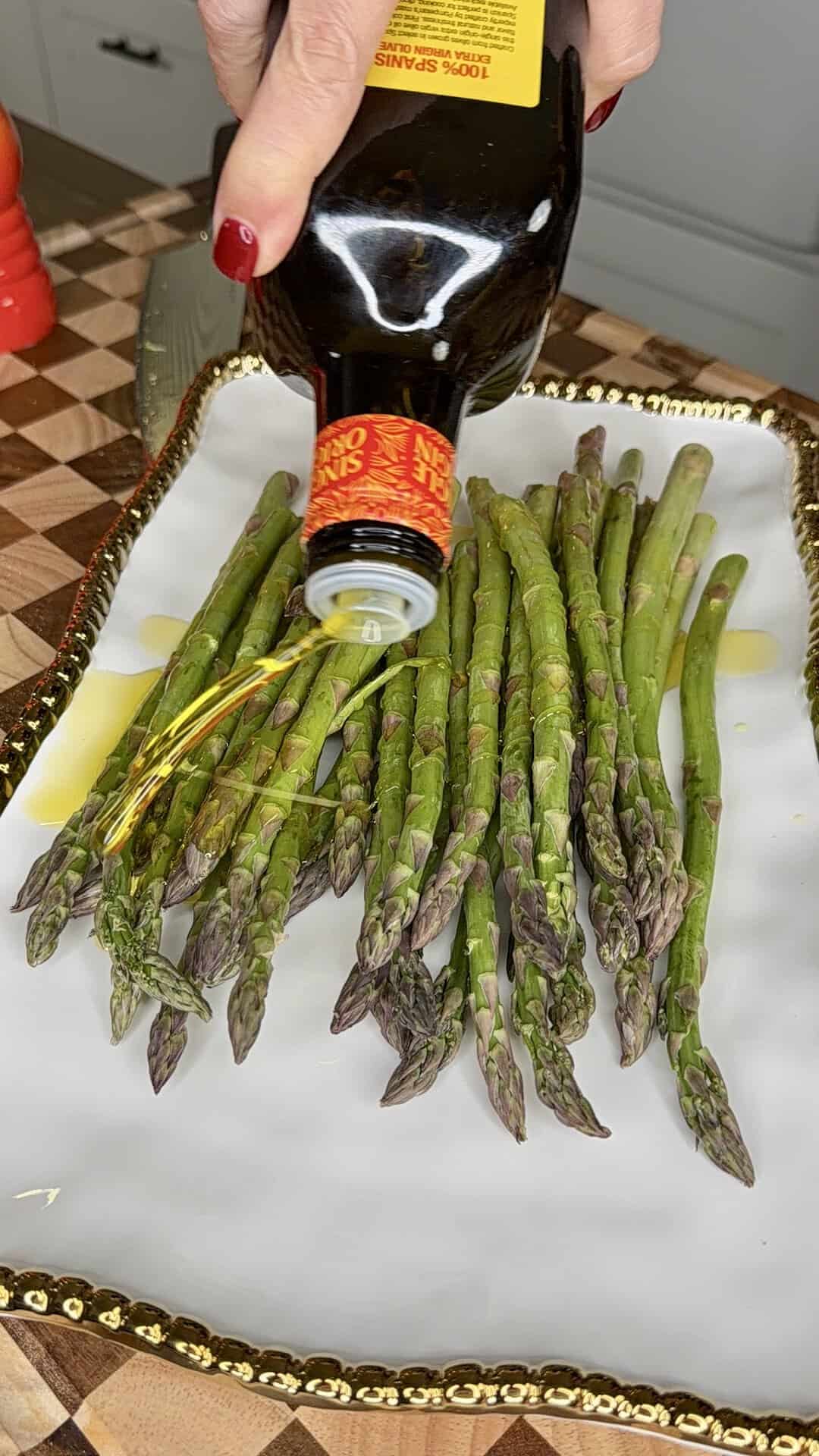 A hand pours olive oil from a bottle onto a pile of fresh asparagus spears arranged on a decorative white and gold platter.