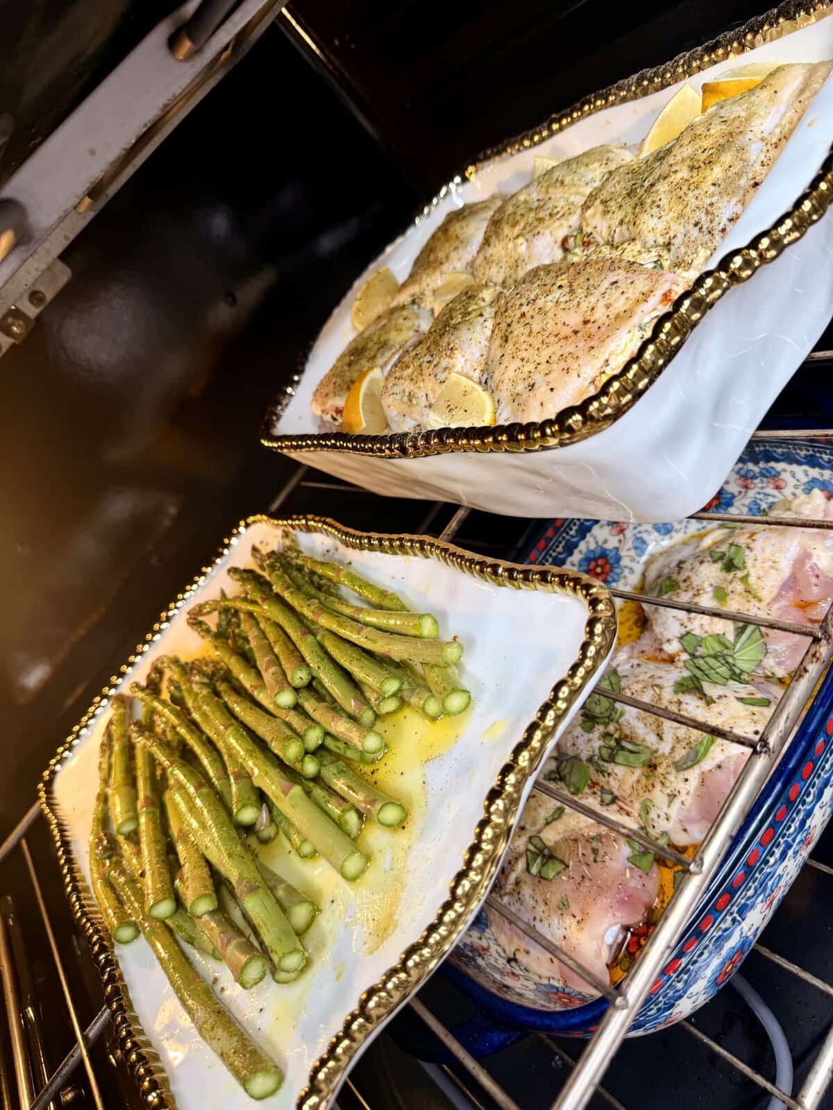 Two white dishes in an oven: one with seasoned chicken and lemon slices, the other with asparagus drizzled in oil. A third colorful dish with more food is partially visible below.
