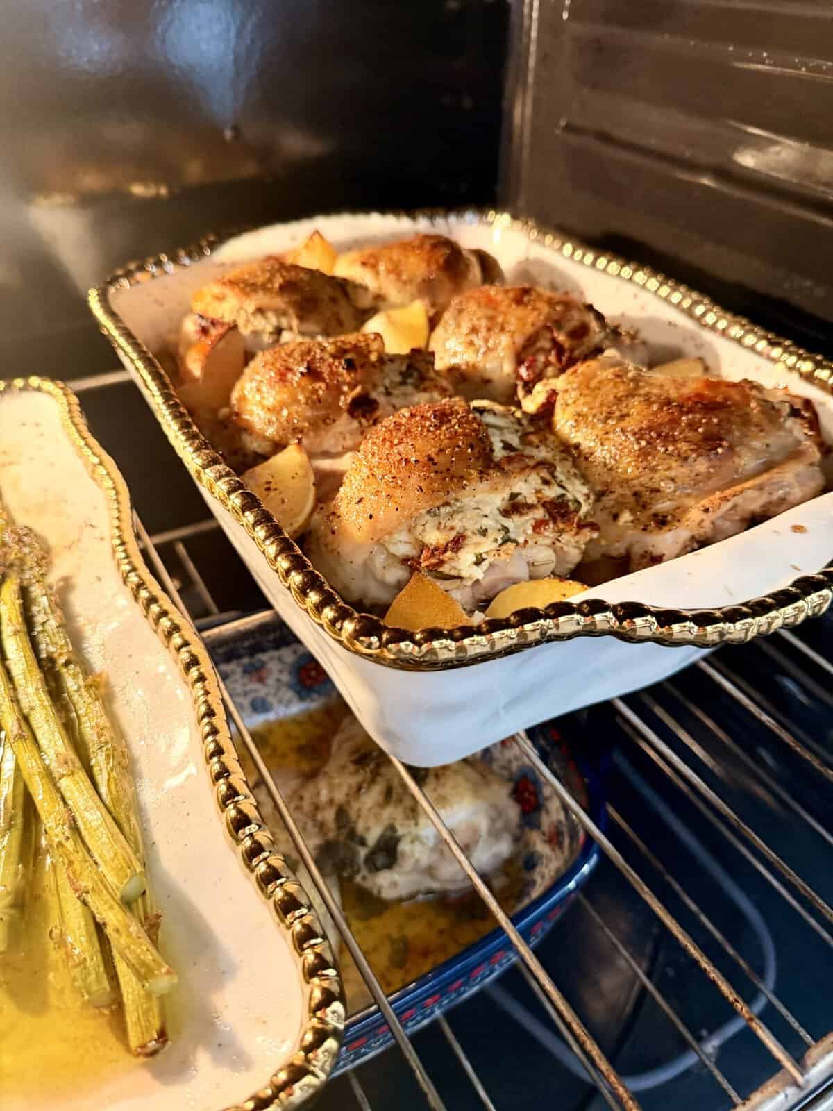 Chicken thighs baked with herbs and lemon slices in a decorative dish, asparagus in a separate dish, and another small dish beneath, all cooking in an oven.