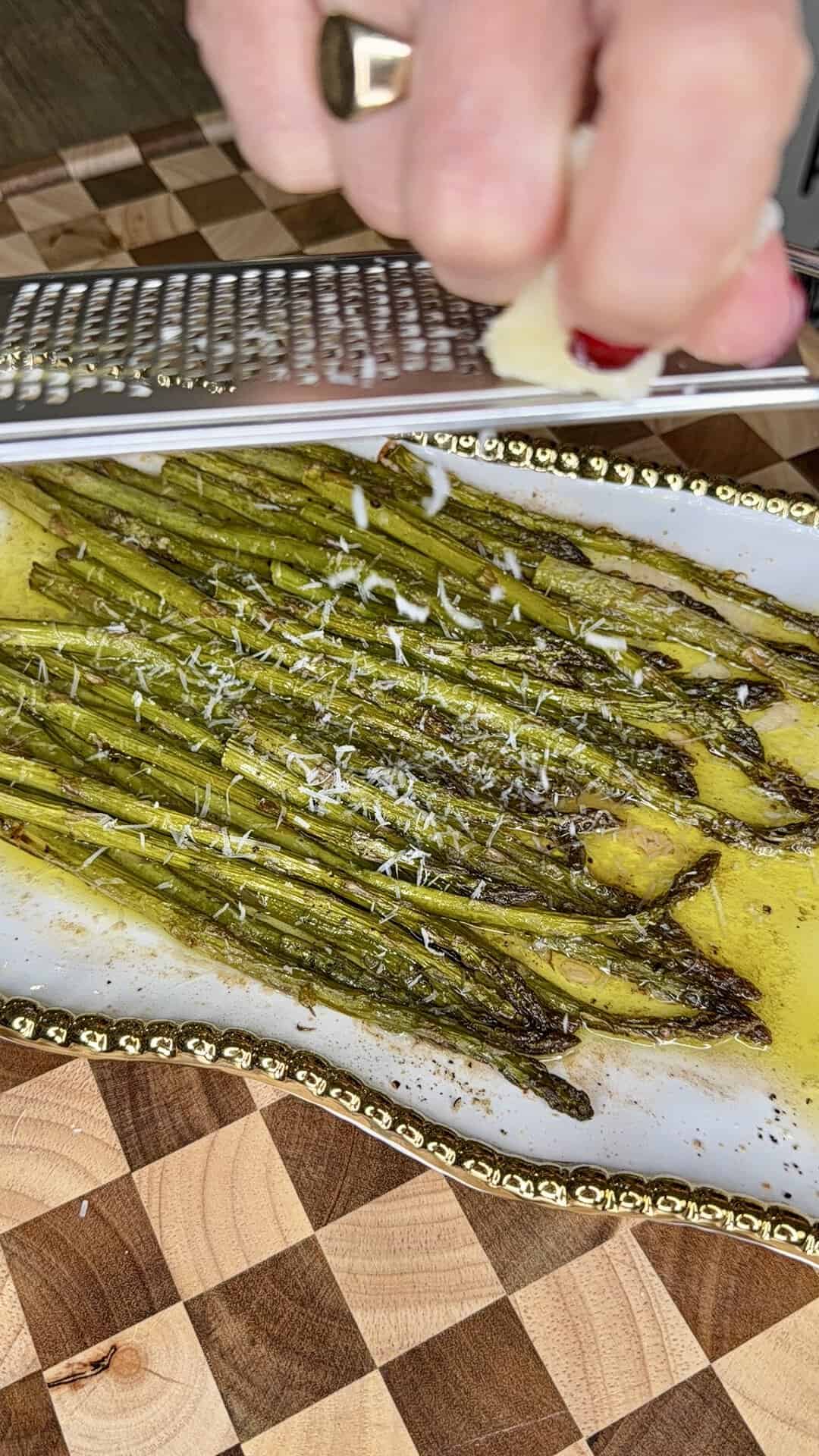A hand grates cheese over roasted asparagus on a white plate with a gold rim, atop a wooden cutting board.