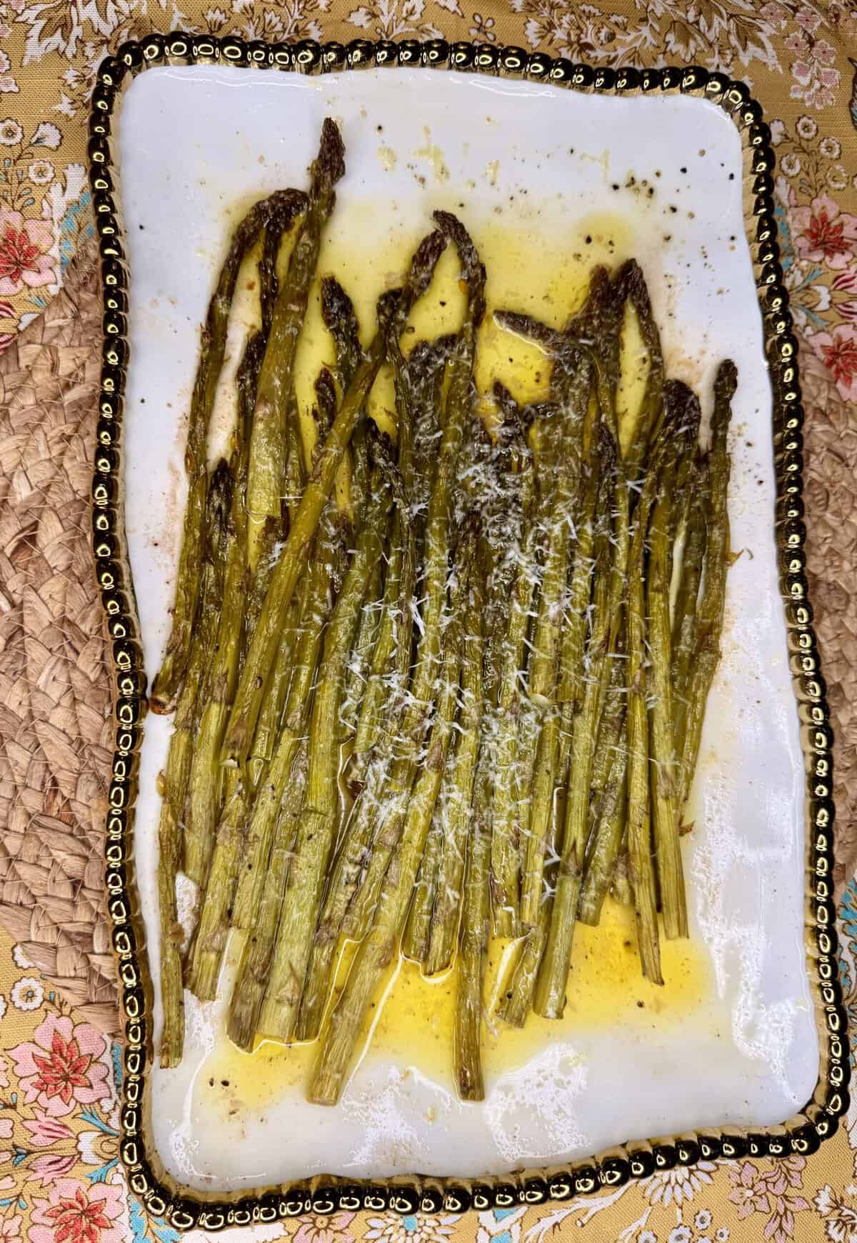 Roasted asparagus spears topped with grated cheese and drizzled with olive oil on a white rectangular plate with a decorative gold beaded edge.