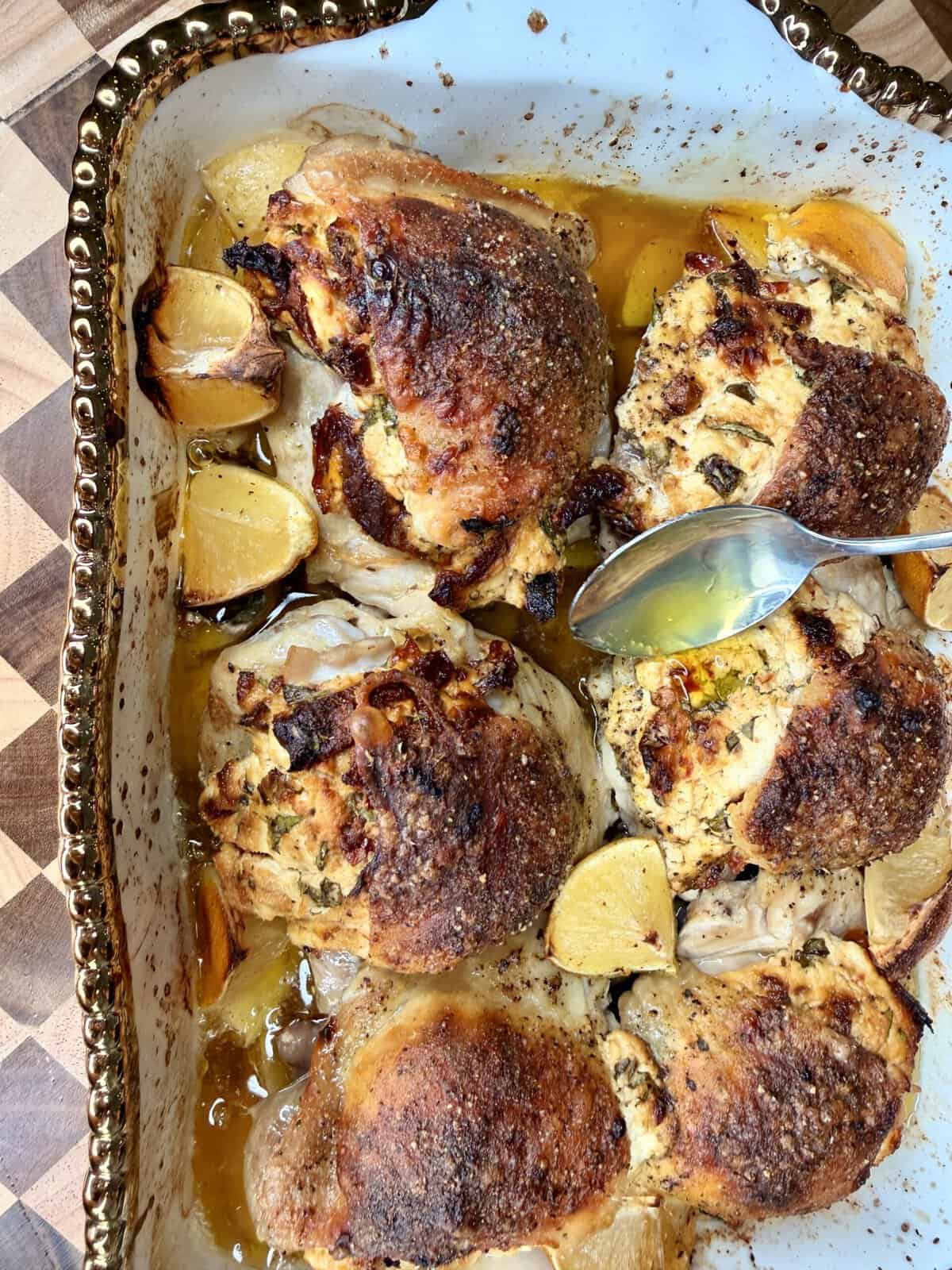 Roast chicken thighs in a ceramic dish with crispy skin, surrounded by lemon wedges and herbs, with a spoon drizzling juices over the meat. The dish sits on a wooden surface.