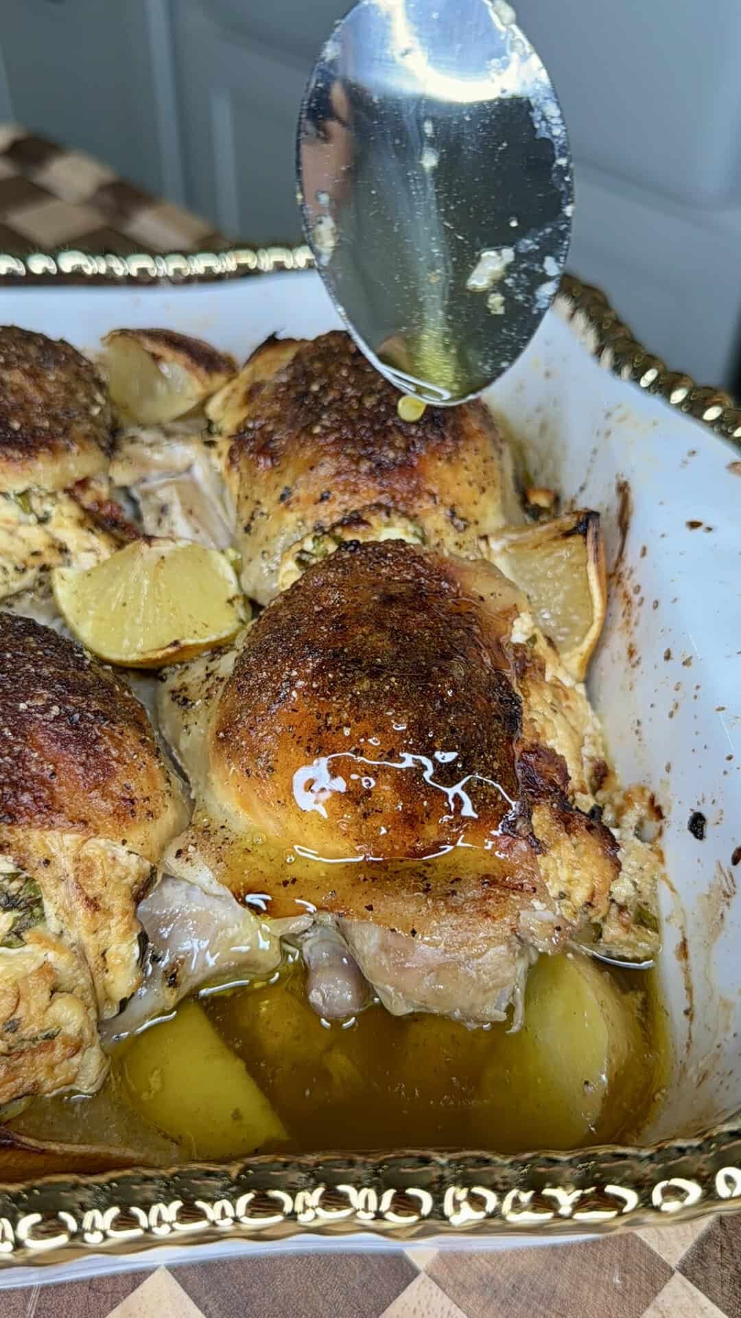A spoon drizzles juices over roasted chicken thighs with crispy skin and lemon wedges in a decorative baking dish.