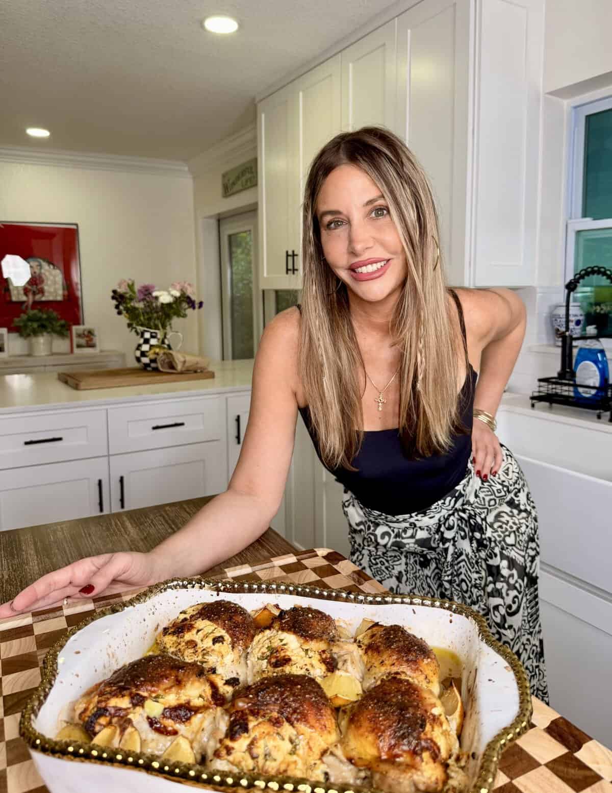 A woman smiles in a kitchen, presenting a baked chicken dish in a decorative pan on a checkered wooden board. White cabinets and flowers are visible in the background.
