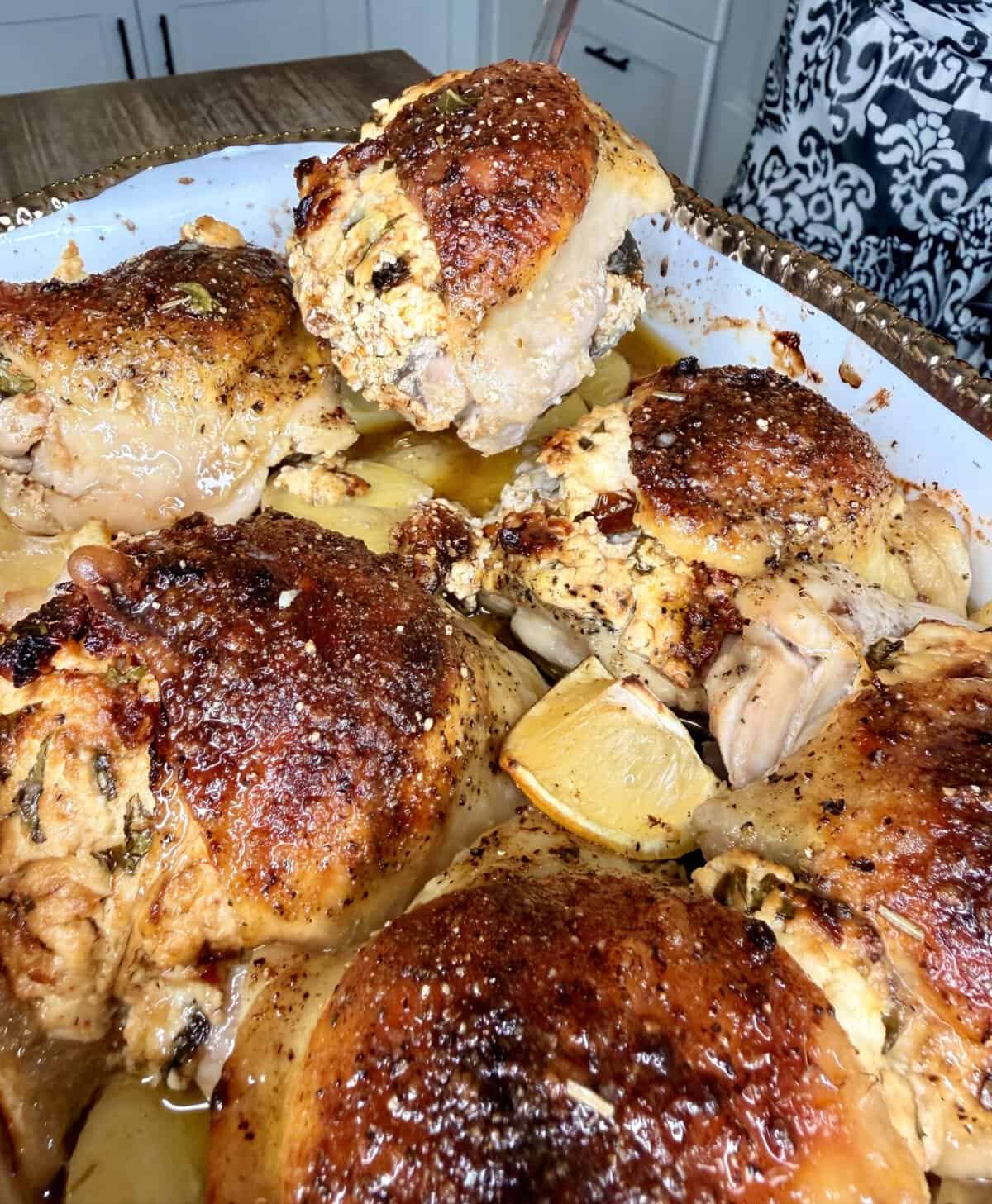 A hand lifts a piece of roasted, golden-brown chicken from a pan filled with juicy baked chicken pieces and lemon wedges, with kitchen cabinets in the background.