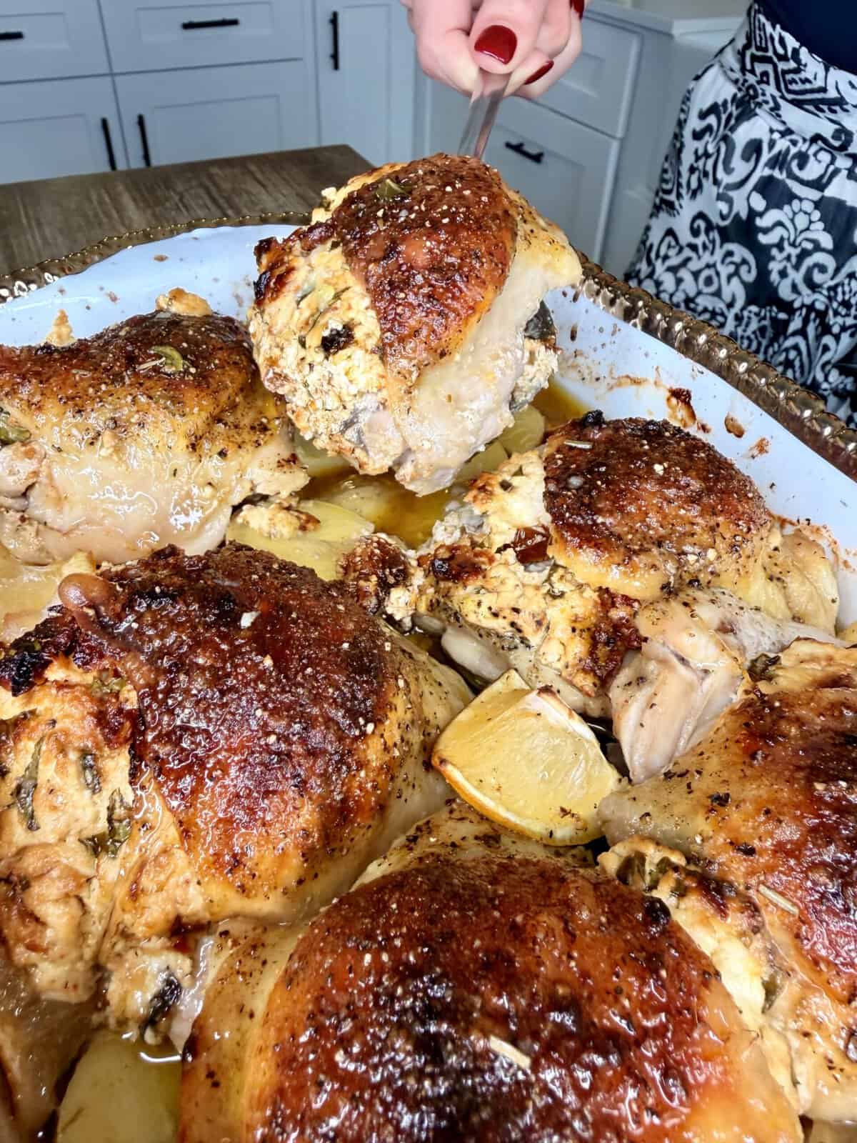 A hand serves roasted chicken pieces with crispy golden skin from a white baking dish, with lemon wedges and herbs visible, on a kitchen counter.