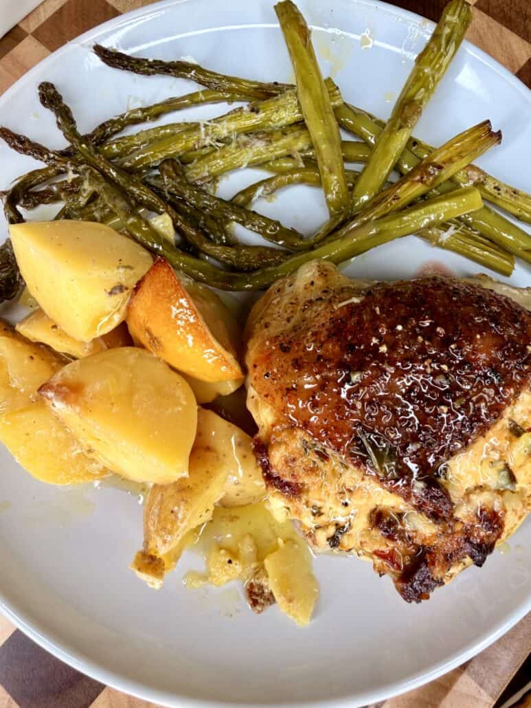 A plate with roasted chicken thigh, golden potatoes, and green asparagus spears, all seasoned and drizzled with sauce, on a white plate atop a wooden surface.