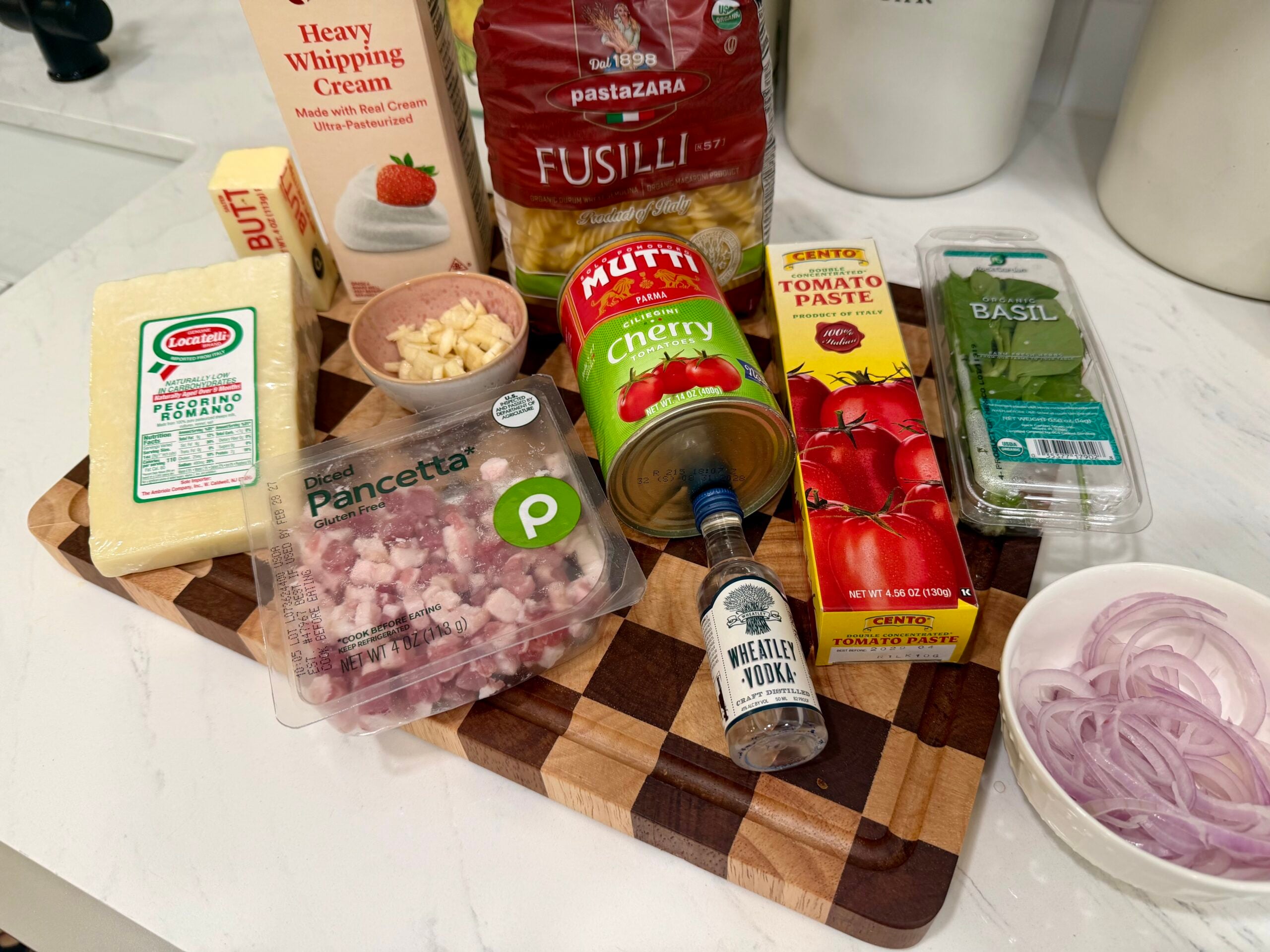 Various pasta ingredients on a cutting board, including fusilli, pancetta, pecorino, tomato paste, cherry tomatoes, basil, chopped garlic, sliced shallots, heavy cream, and a small bottle of vodka.