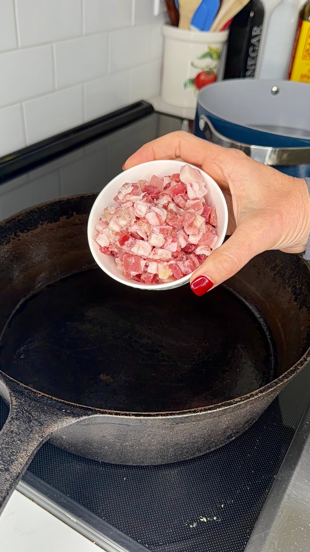 A hand with red nail polish holds a white bowl of frozen, diced meat over a black cast iron skillet on a stove.