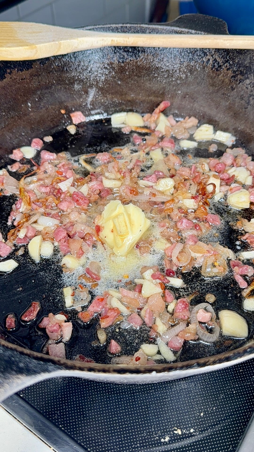 Chopped garlic, onions, and bacon are being sautéed with a pat of butter in a cast iron skillet on a stovetop.