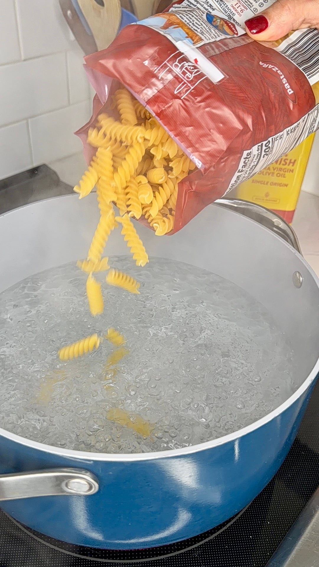 A hand pours rotini pasta from a red bag into a blue pot of boiling water on a stovetop. Steam rises from the pot.