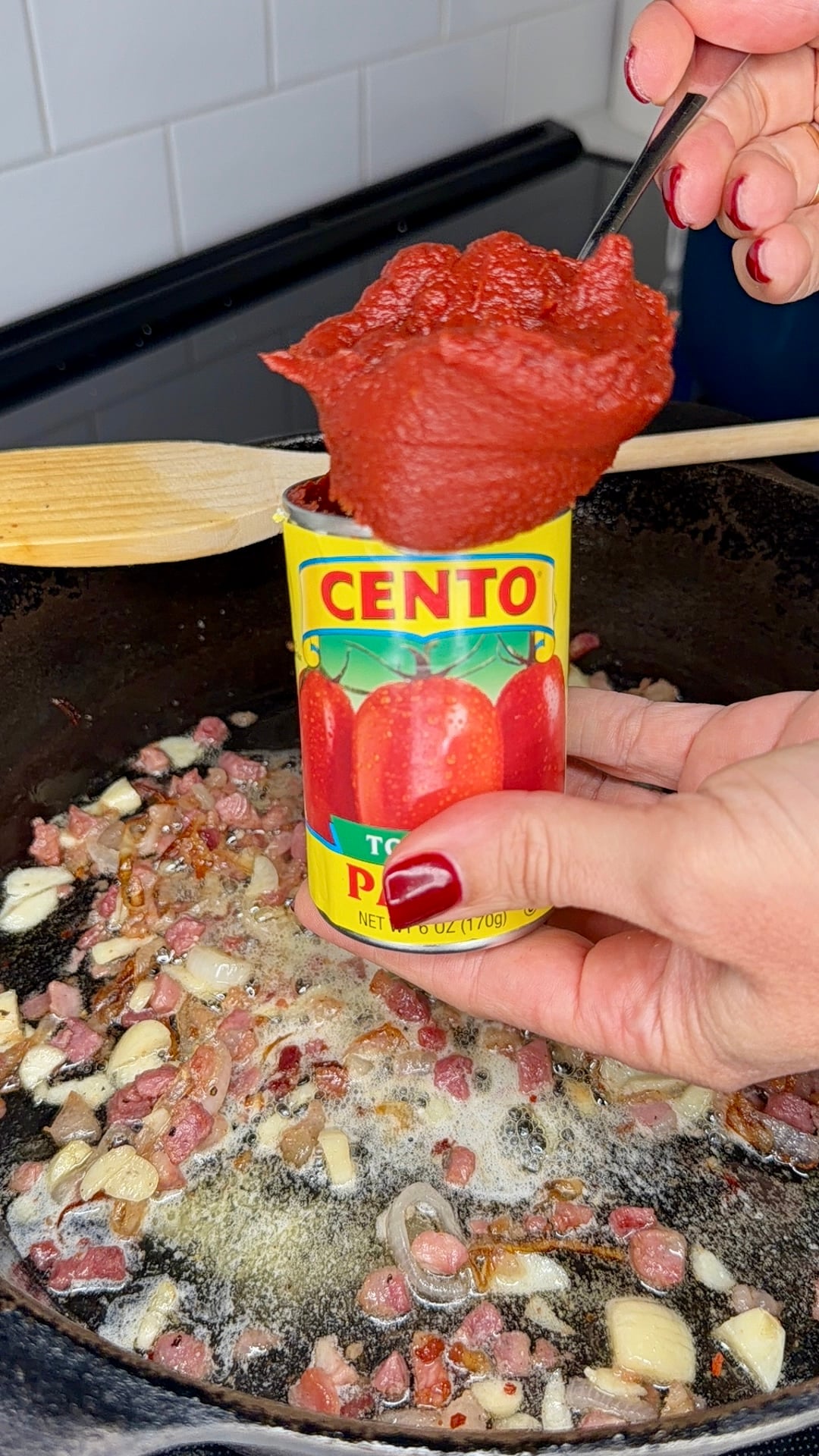 A hand scoops tomato paste from a Cento can over a skillet with sautéing pancetta, garlic, and onions.