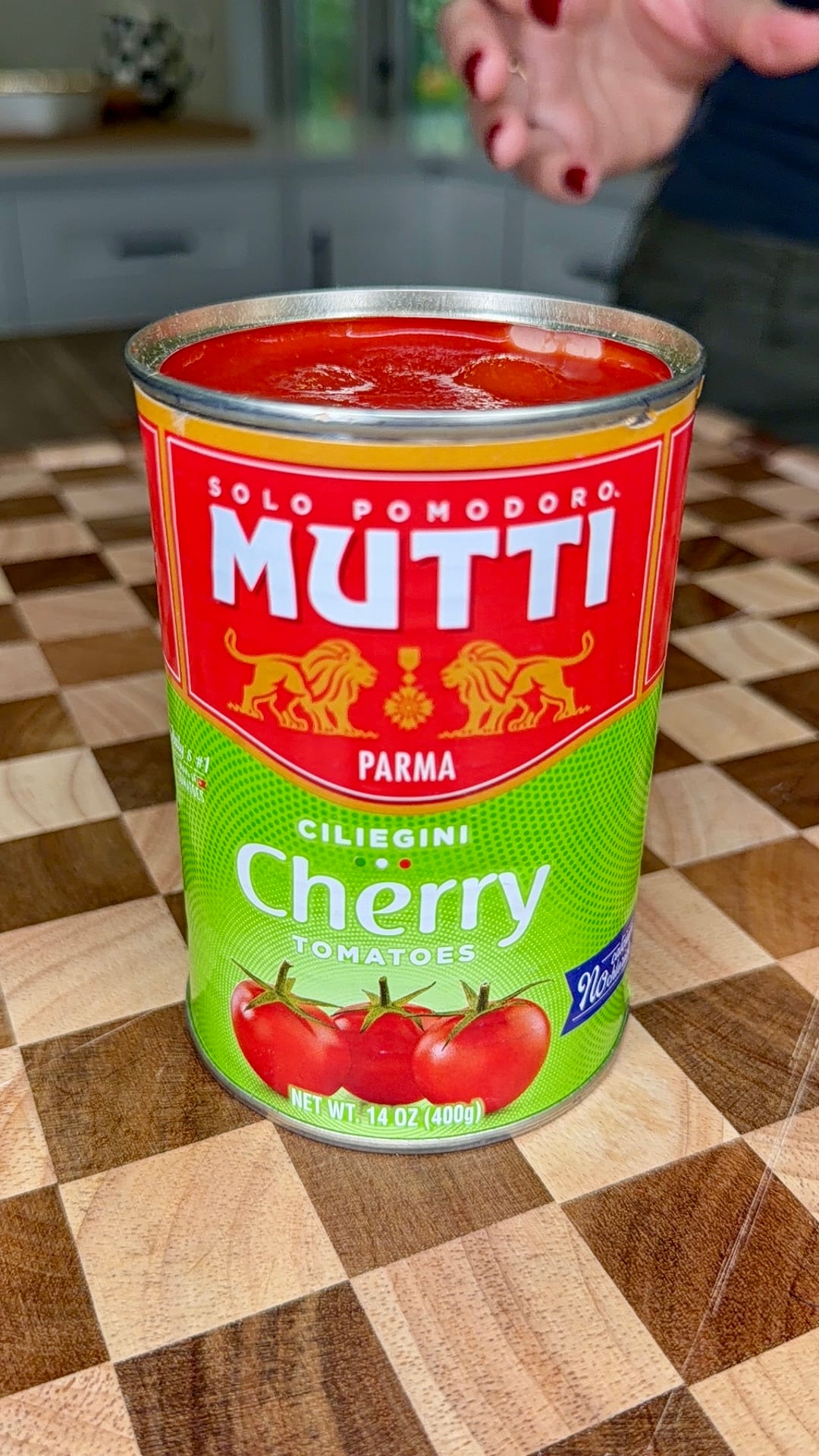A can of Mutti Cherry Tomatoes sits on a checkered wooden surface. The can is open, showing the tomatoes inside. A hand with red-painted nails appears in the background.