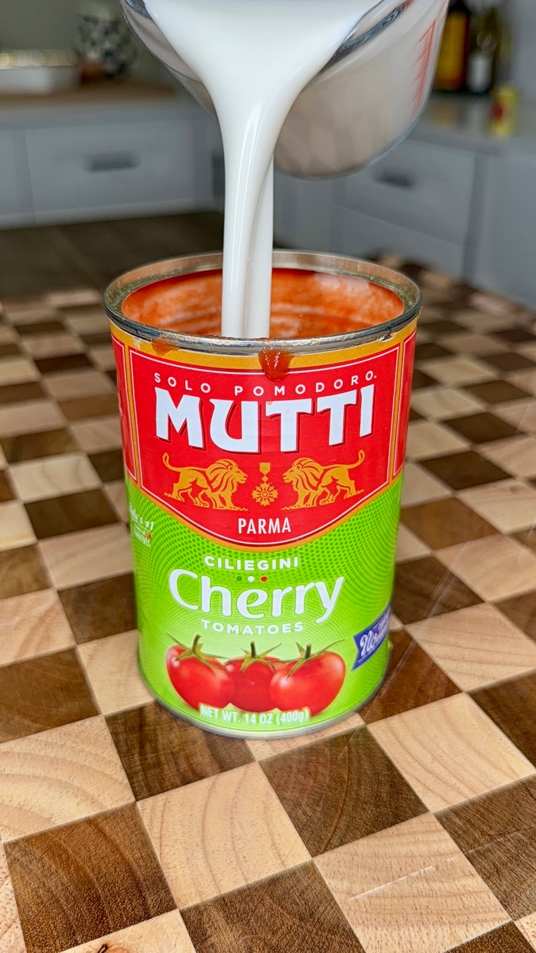 A can of Mutti cherry tomatoes sits on a wooden checkered surface as a white liquid is being poured into it from a measuring cup in a kitchen setting.
