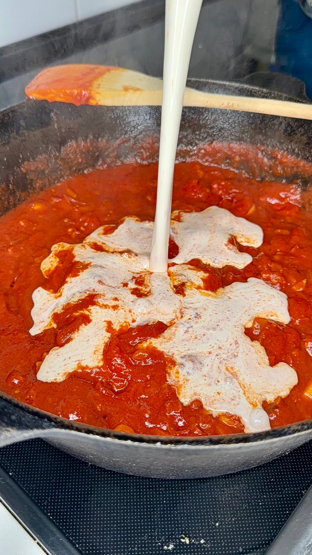 Cream is being poured into a skillet of red tomato sauce on a stovetop, with a wooden spoon resting on the side of the pan.