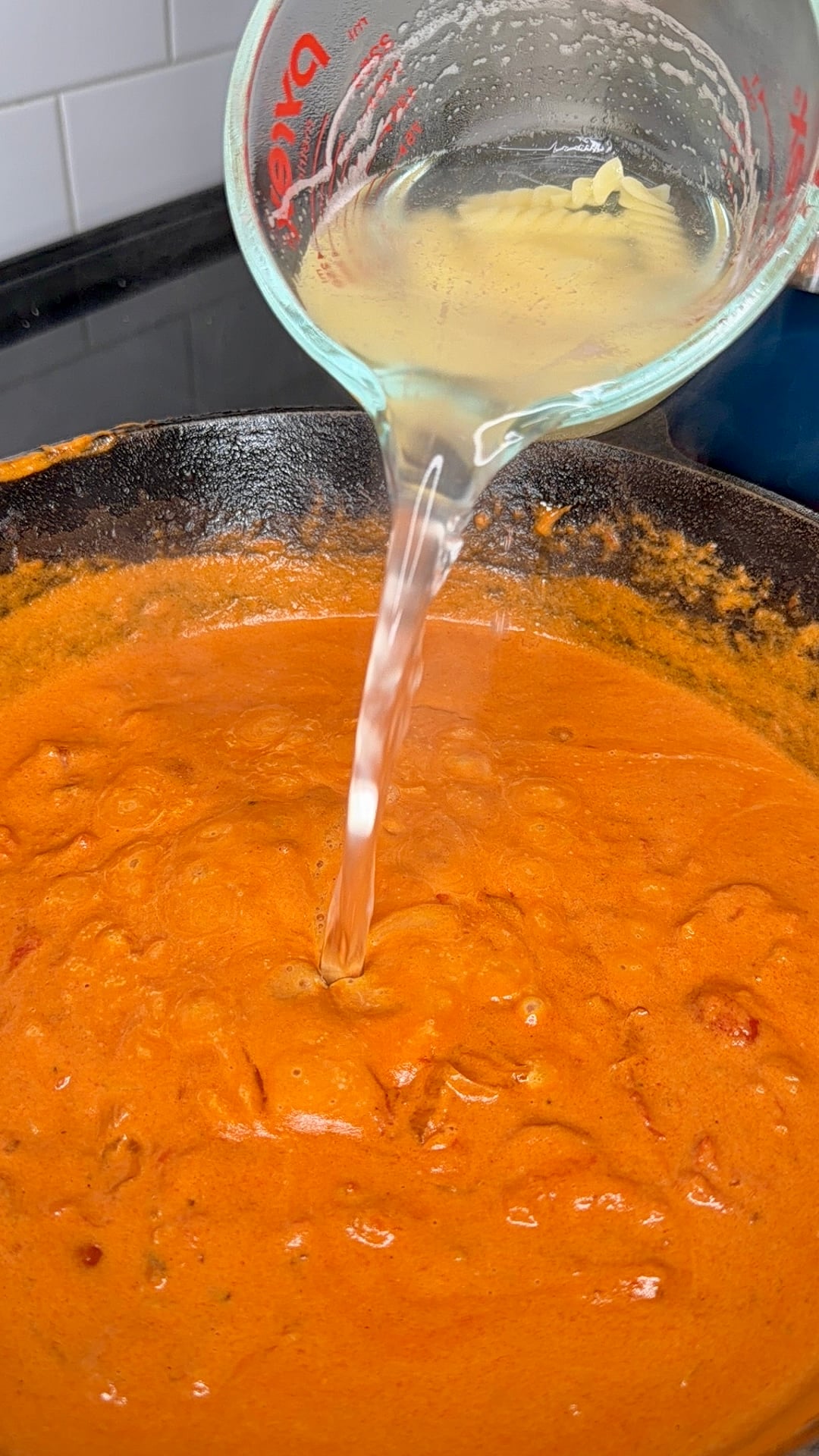 A glass measuring cup is pouring liquid into a simmering orange-red sauce in a black pan on a stove.
