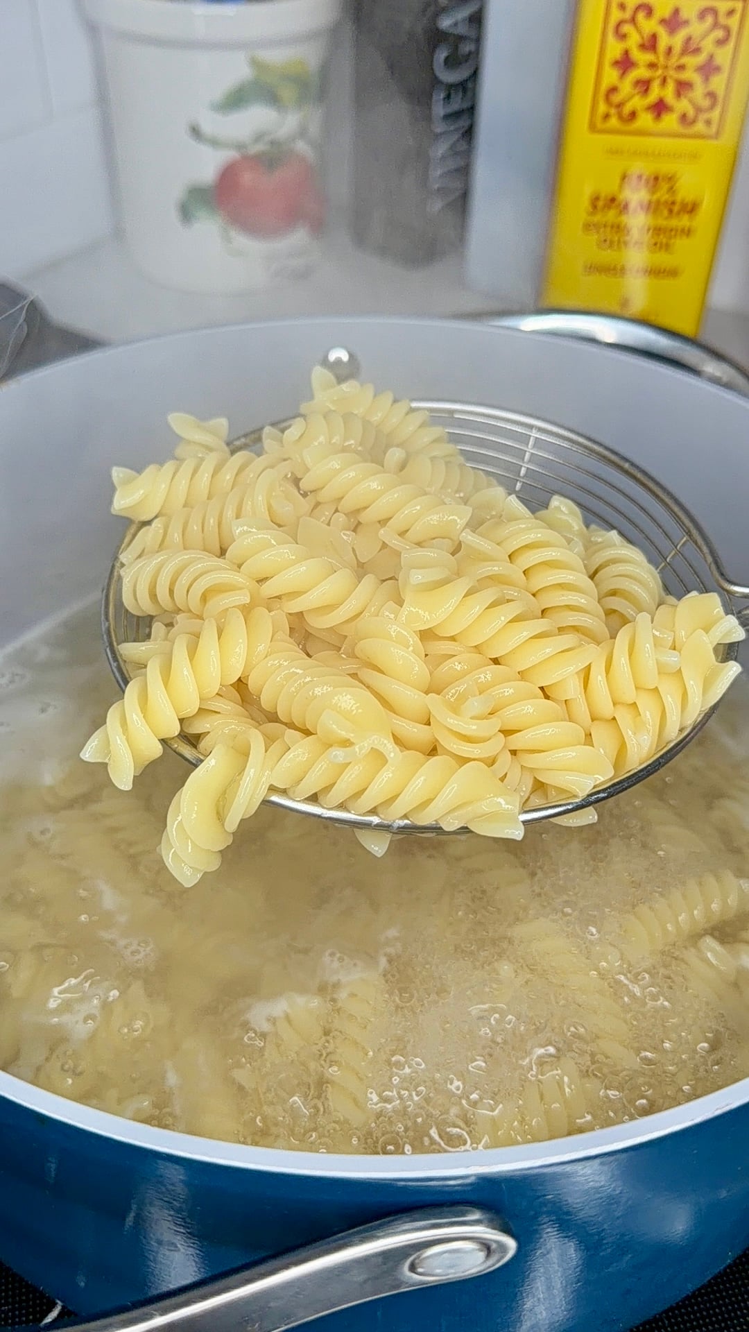 A wire skimmer lifts cooked fusilli pasta from a pot of boiling water. In the background, a tomato-patterned canister and a bottle of olive oil sit on the counter.