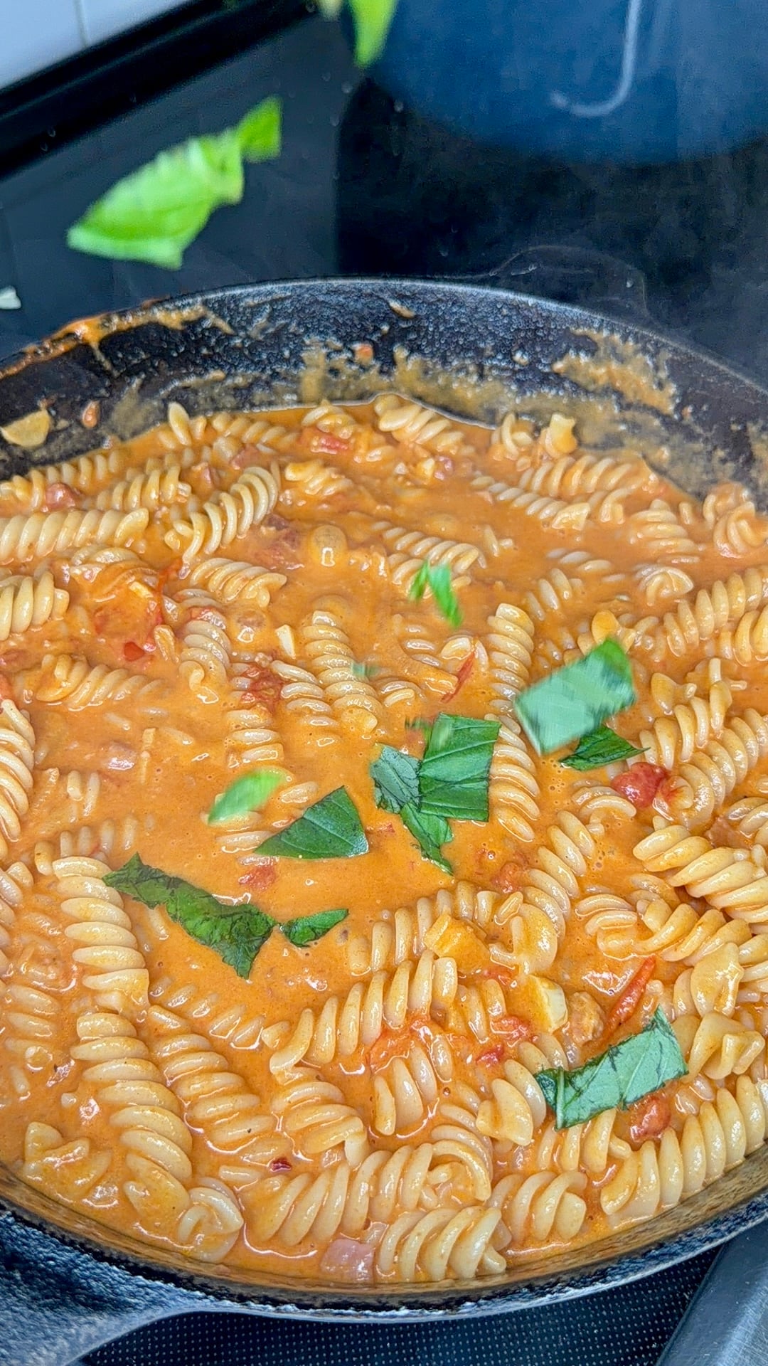 A skillet filled with creamy tomato sauce fusilli pasta, garnished with fresh basil leaves, sits on a stovetop.