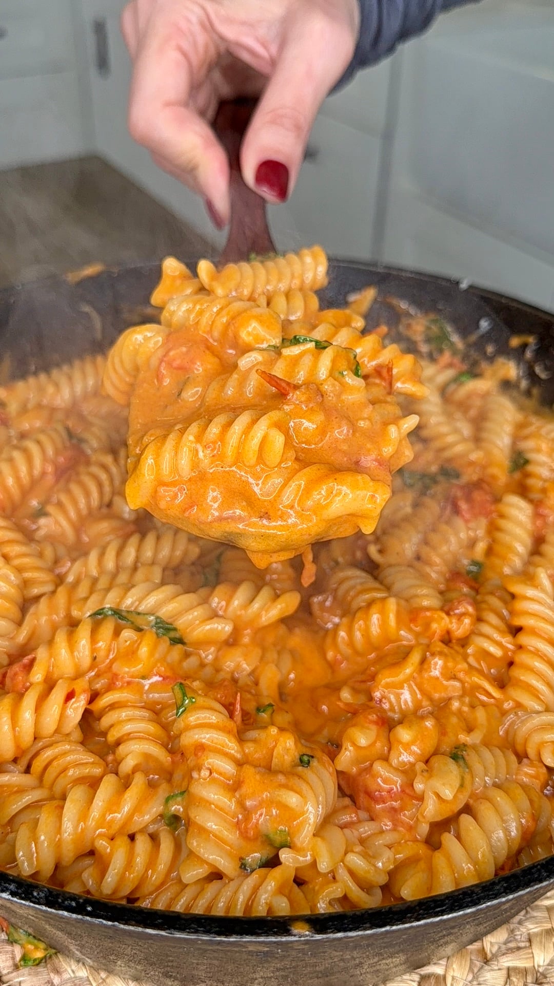 A hand with red nail polish holds a serving of creamy tomato rotini pasta above a pan filled with the same pasta, topped with fresh herbs. Steam rises from the hot dish.